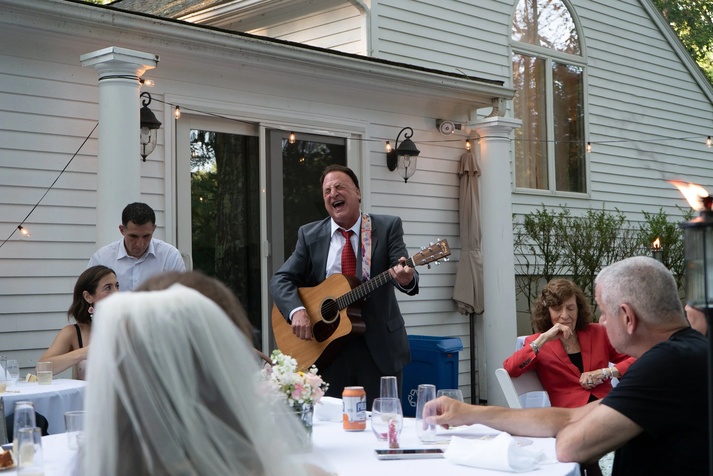 Man in a suit singing and playing guitar at an outdoor wedding reception.