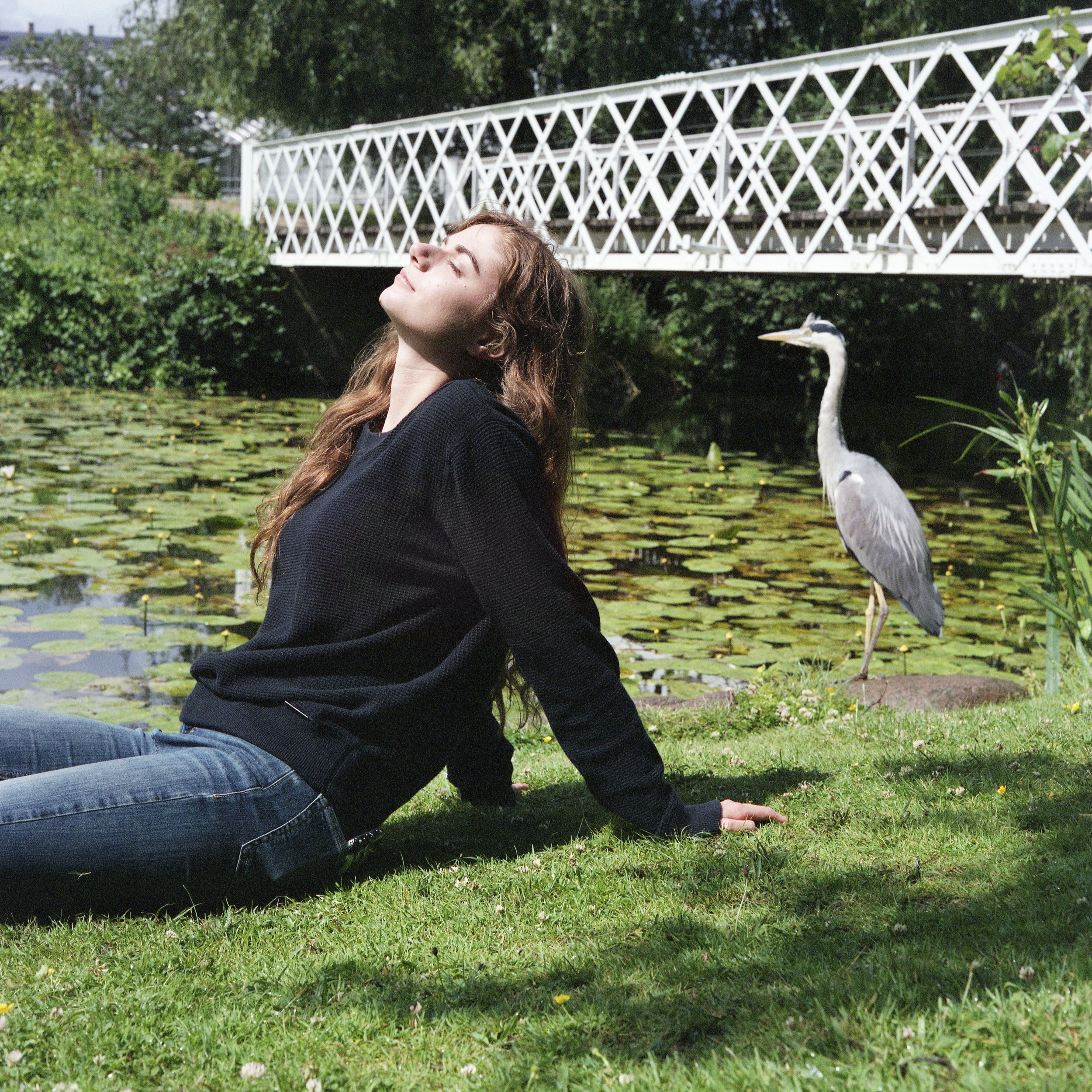 A young woman with long brown hair sitting on green grass near a pond with lily pads, her head tilted back and eyes closed, in sunny weather with a white bridge and a heron in the background.