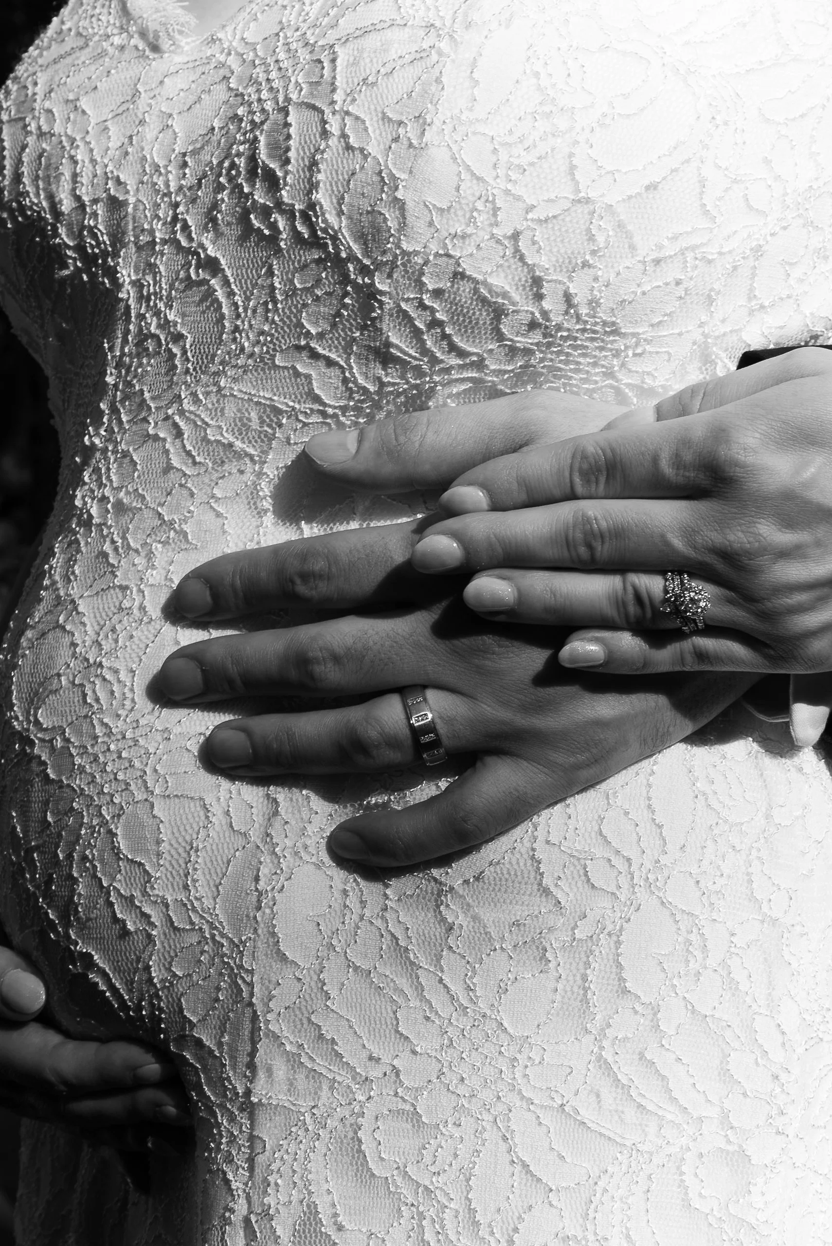 Close-up of a person in bridal attire with hands resting on their torso, showing wedding rings and a lace dress.