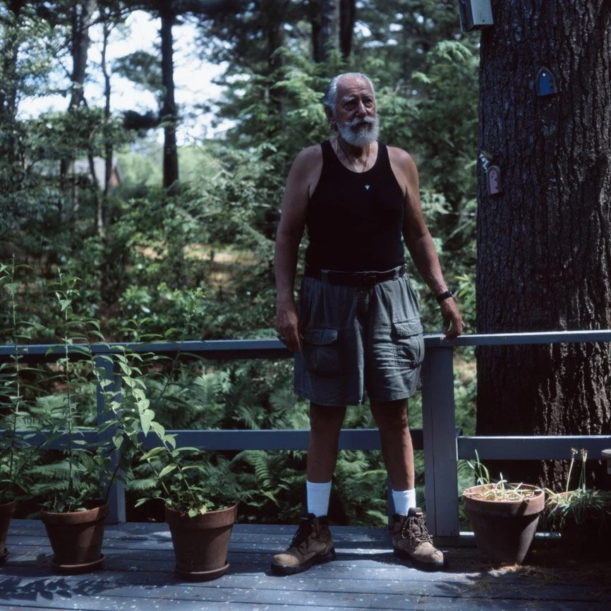 An elderly man with a white beard, wearing a black tank top, gray shorts, hiking boots, and white socks, standing on a wooden deck outdoors surrounded by greenery and potted plants, next to a large tree with trail camera mounted on it.