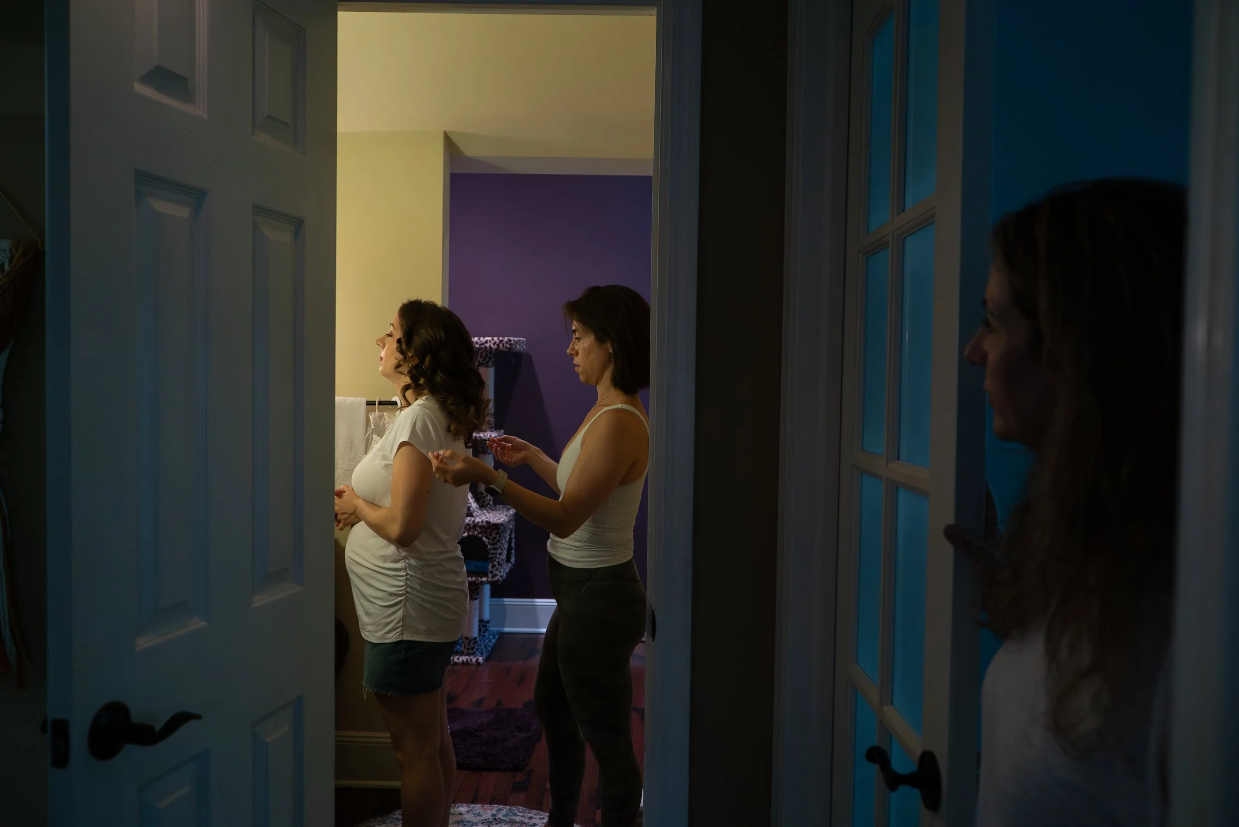 Three women in a bedroom, one adjusting her clothes, another applying makeup, and the third peeking from a doorway, with purple walls and a leopard print chair in the background.