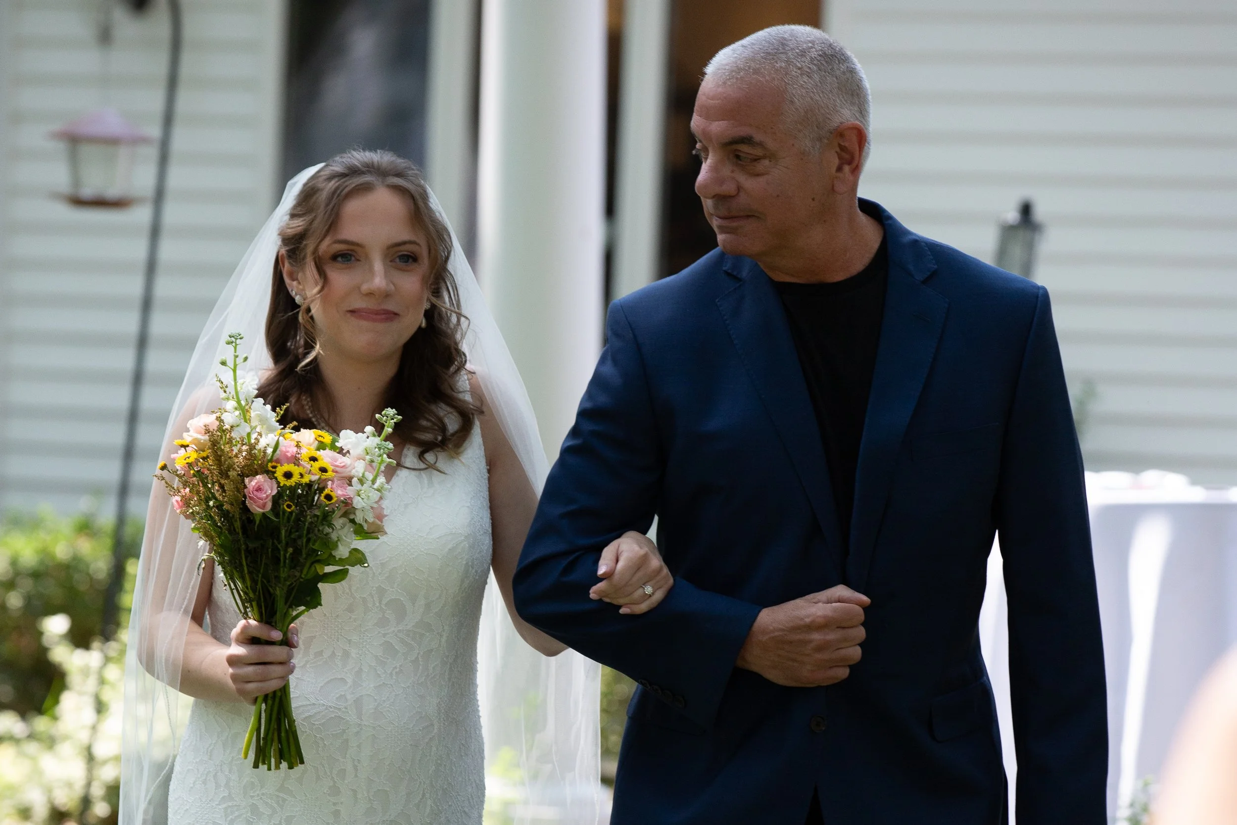 A bride in a white wedding dress holding a bouquet of flowers, walking arm-in-arm with a man in a navy blue suit at an outdoor wedding ceremony.