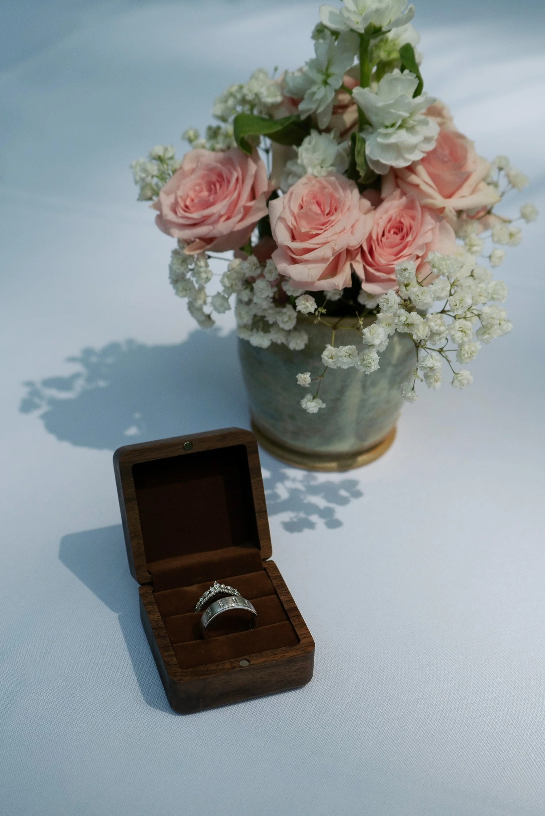A bouquet of pink roses and white baby's breath in a ceramic vase, with a wooden ring box containing a wedding ring and engagement ring on a white surface.