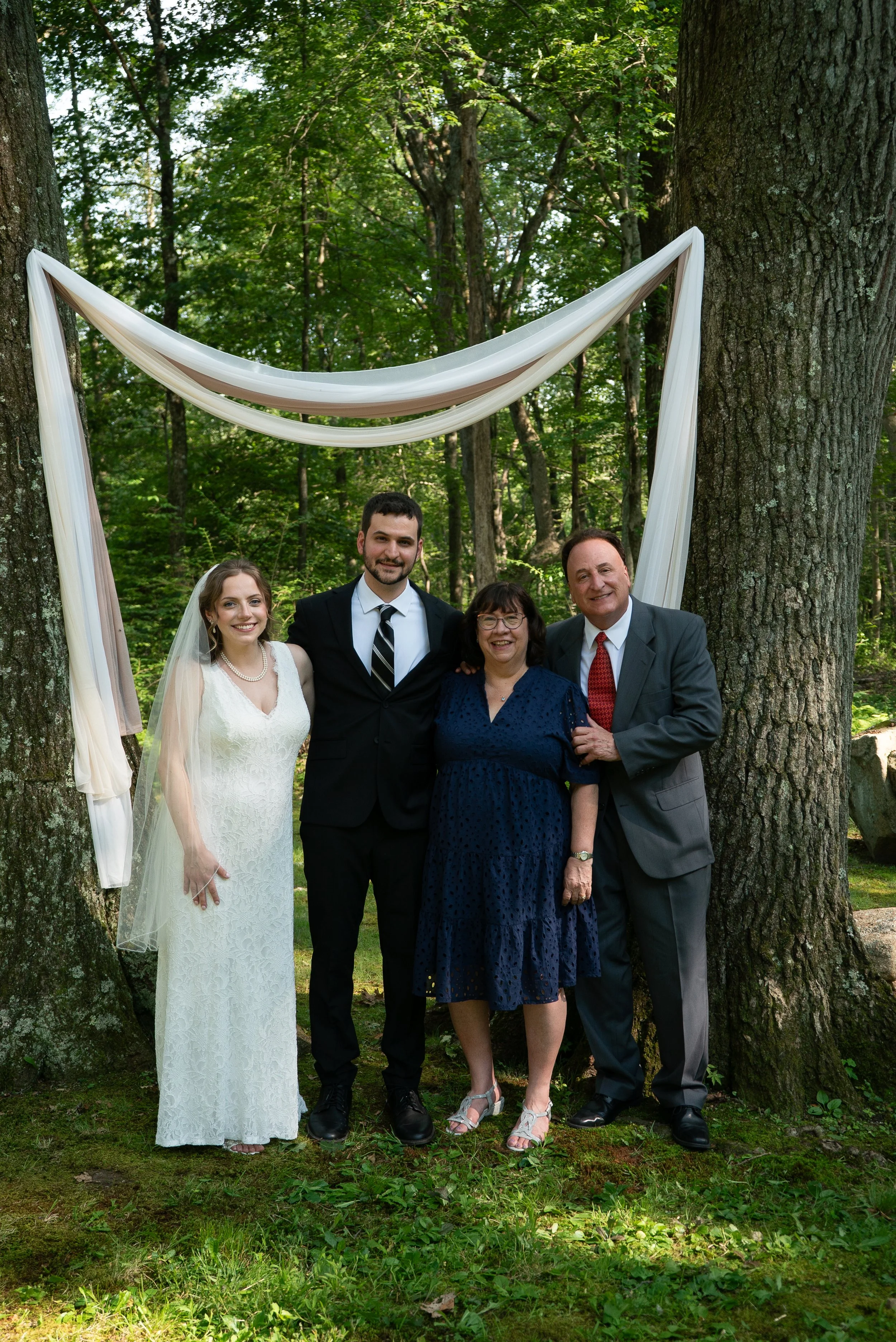Group of five people standing outdoors in a wooded area, dressed for a wedding, posing under a fabric-draped arch.
