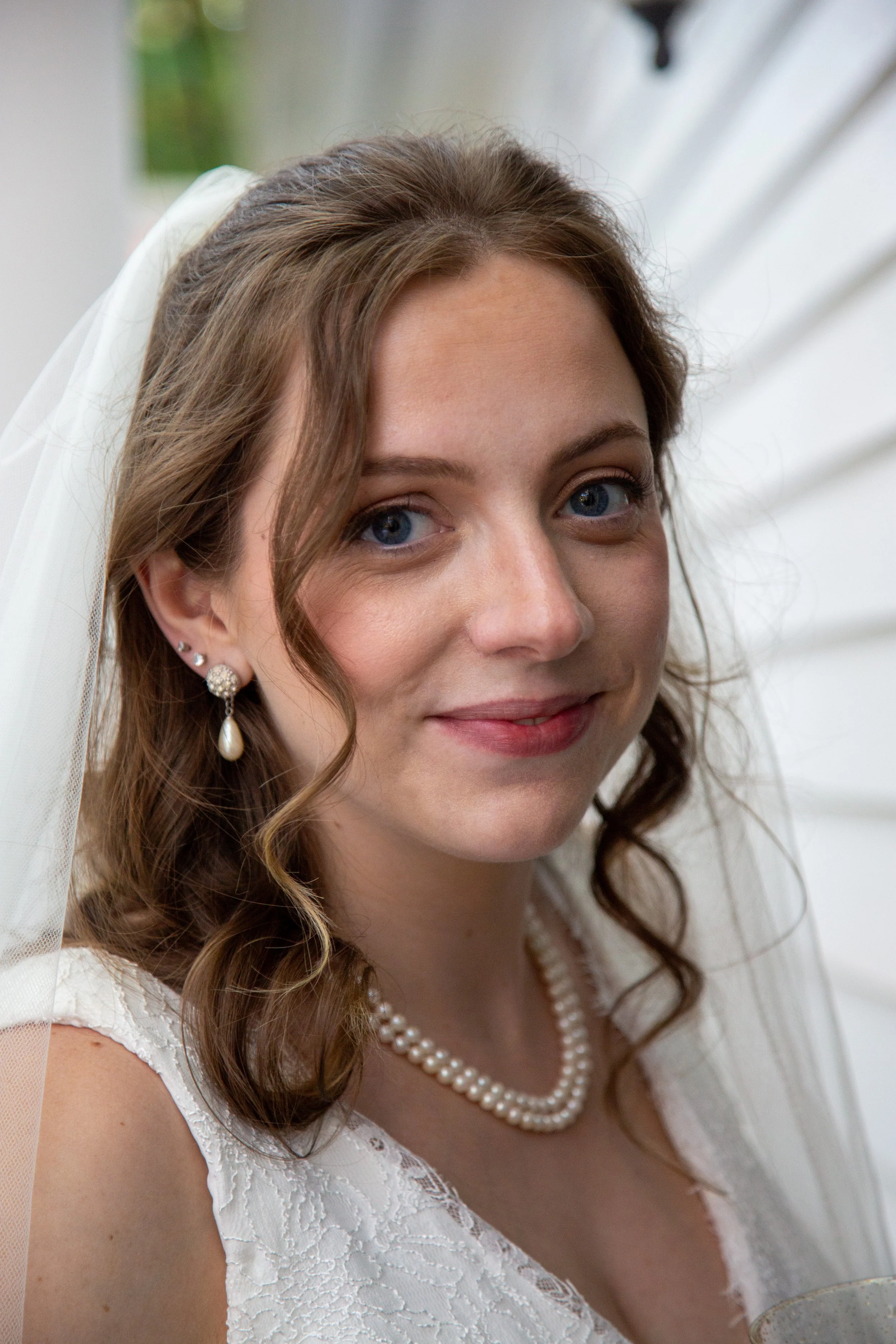 A bride with brown, curly hair, blue eyes, wearing pearl earrings, a pearl necklace, and a white lace dress, smiling with a white veil and soft background.