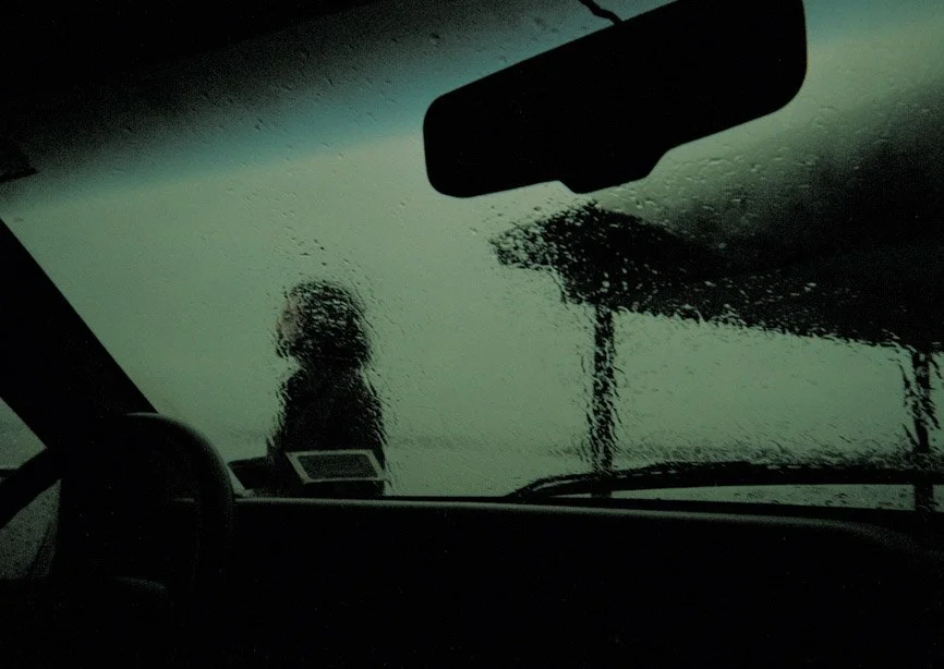 Interior of a vehicle during heavy rain, with a view of rain-streaked windshield, rearview mirror, and a silhouette of a person outside.
