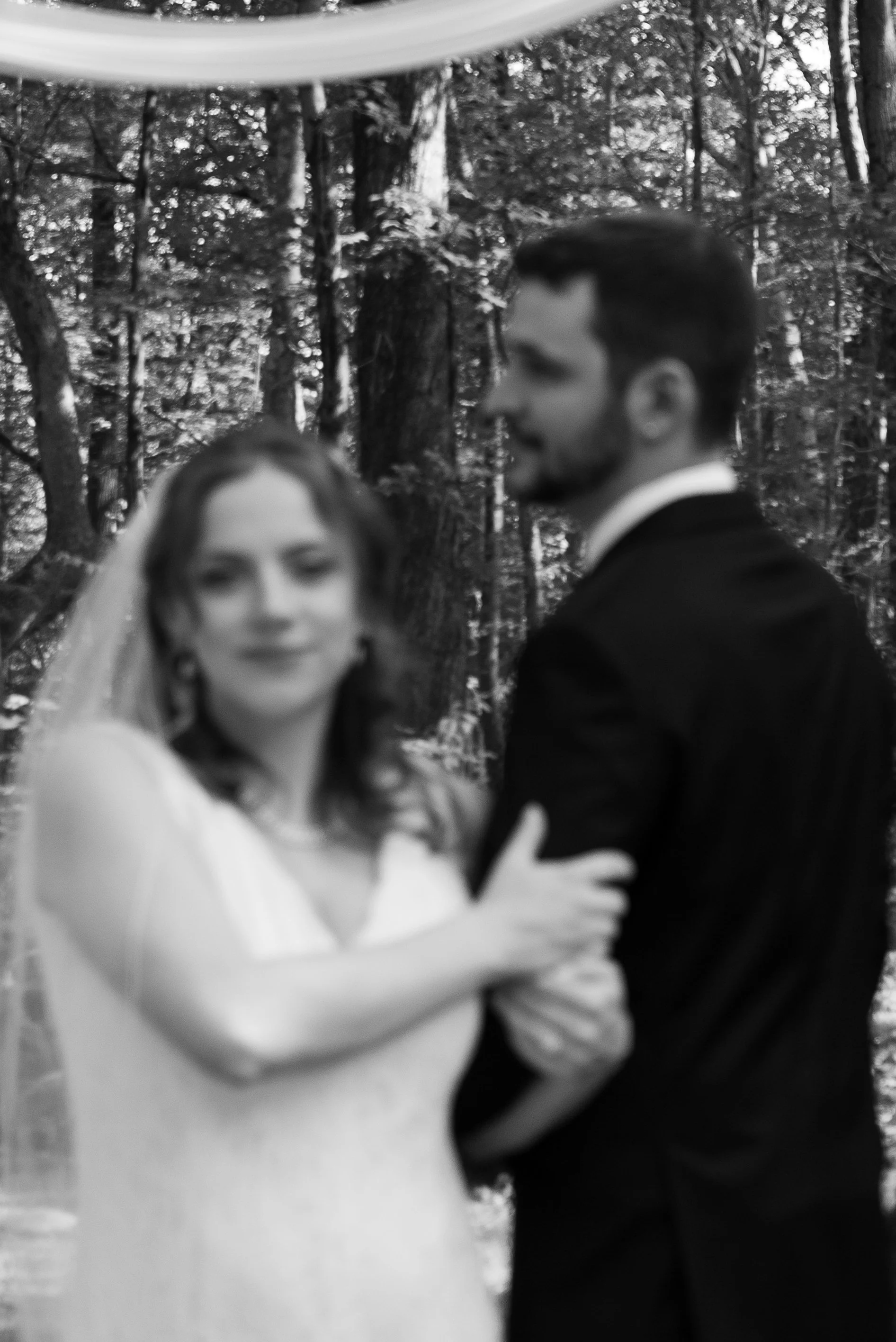 Blurred black-and-white photo of a bride and groom standing closely together outdoors in a wooded area, with the bride smiling and the groom looking to the side.