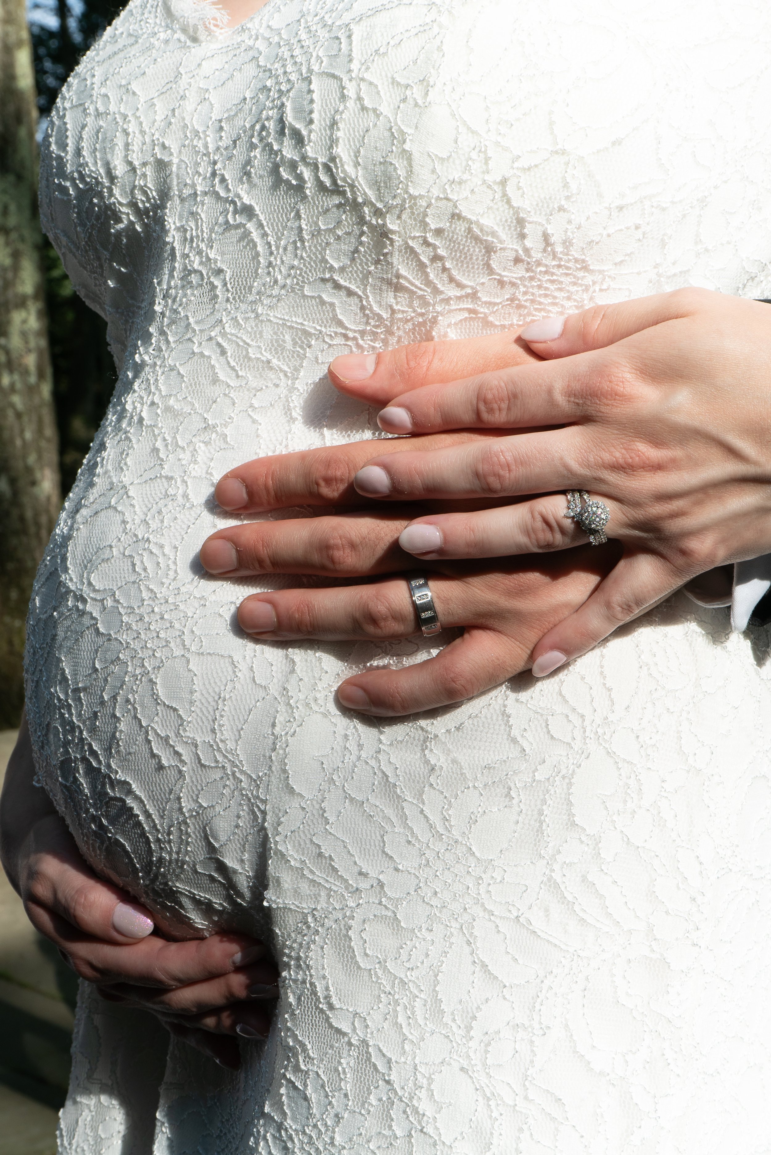 Close-up of a pregnant woman in a white lace dress with hands on her belly, showing wedding and engagement rings.