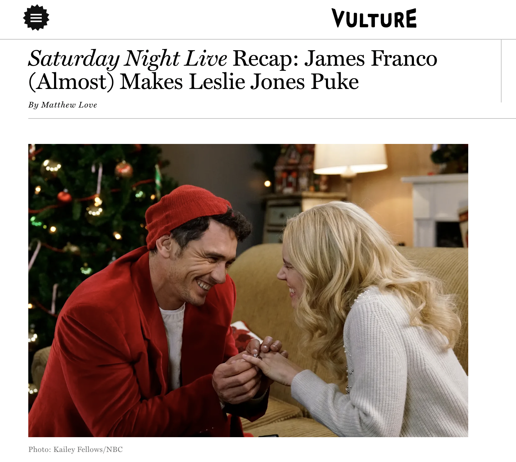 A man dressed in a red jacket and Santa hat proposes marriage to a woman with blonde hair, both smiling, in a living room decorated for Christmas with a tree in the background.