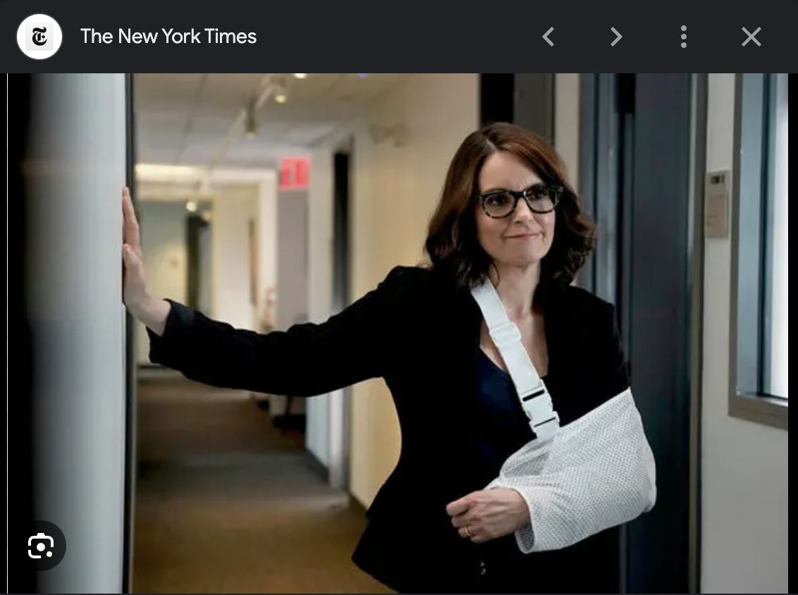 A woman with shoulder-length brown hair, glasses, and a black blazer stands in a hallway with her arm in a sling. She is smiling and has her hand resting on a wall.