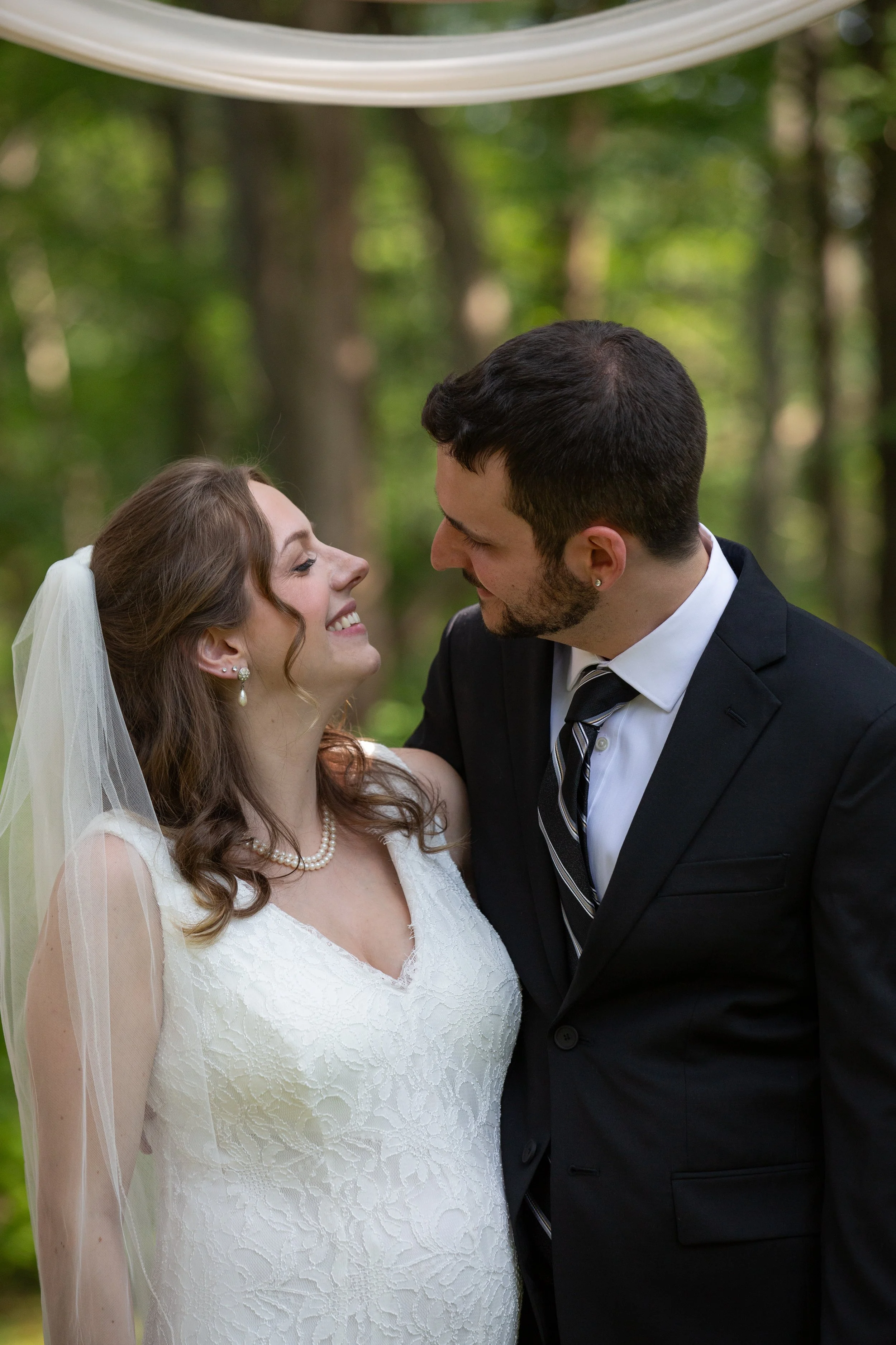 A newlywed couple sharing an intimate moment in a lush outdoor setting, with the bride wearing a white lace wedding dress and veil, and the groom in a black suit and tie.