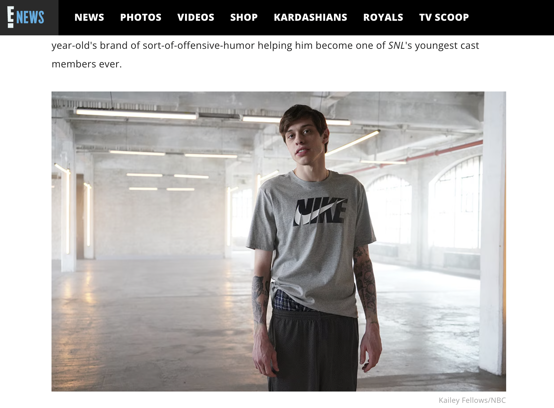 Young man with tattoos wearing a gray Nike t-shirt, standing in a spacious, industrial-style room with large windows and exposed ceiling pipes.