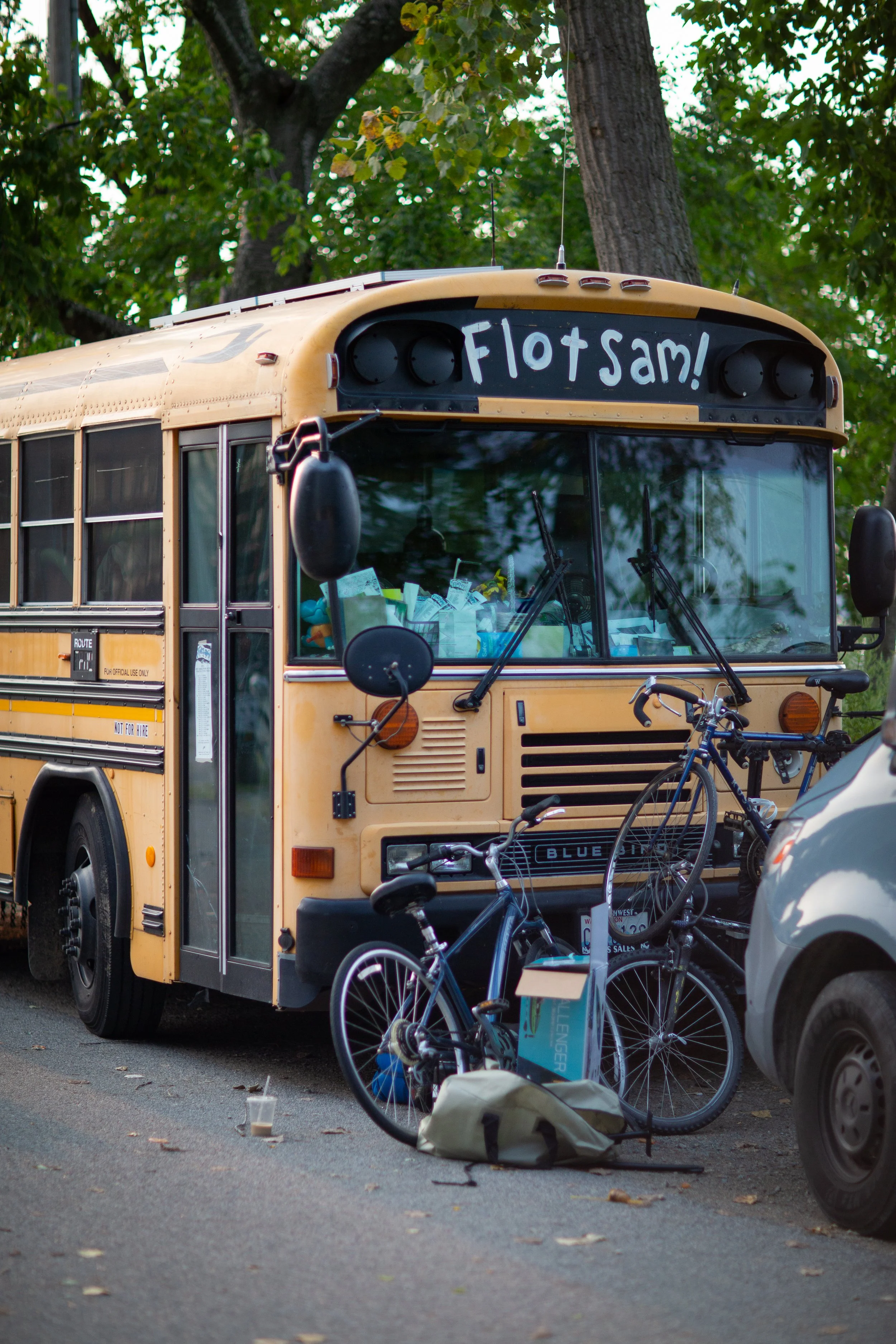 Yellow school bus with a sign reading 'Flotsam!' on the front, parked on a street next to two bicycles and a gray van. Behind the bus are trees with green leaves.