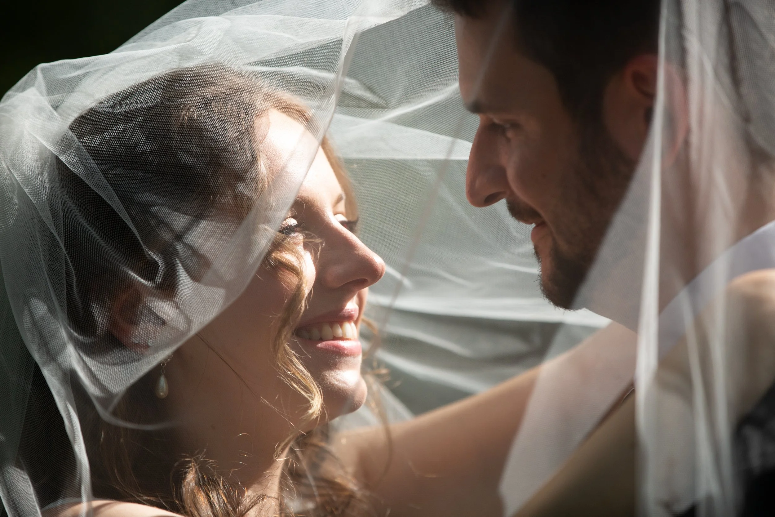 A bride and groom smiling at each other under a sheer veil in an outdoor setting.