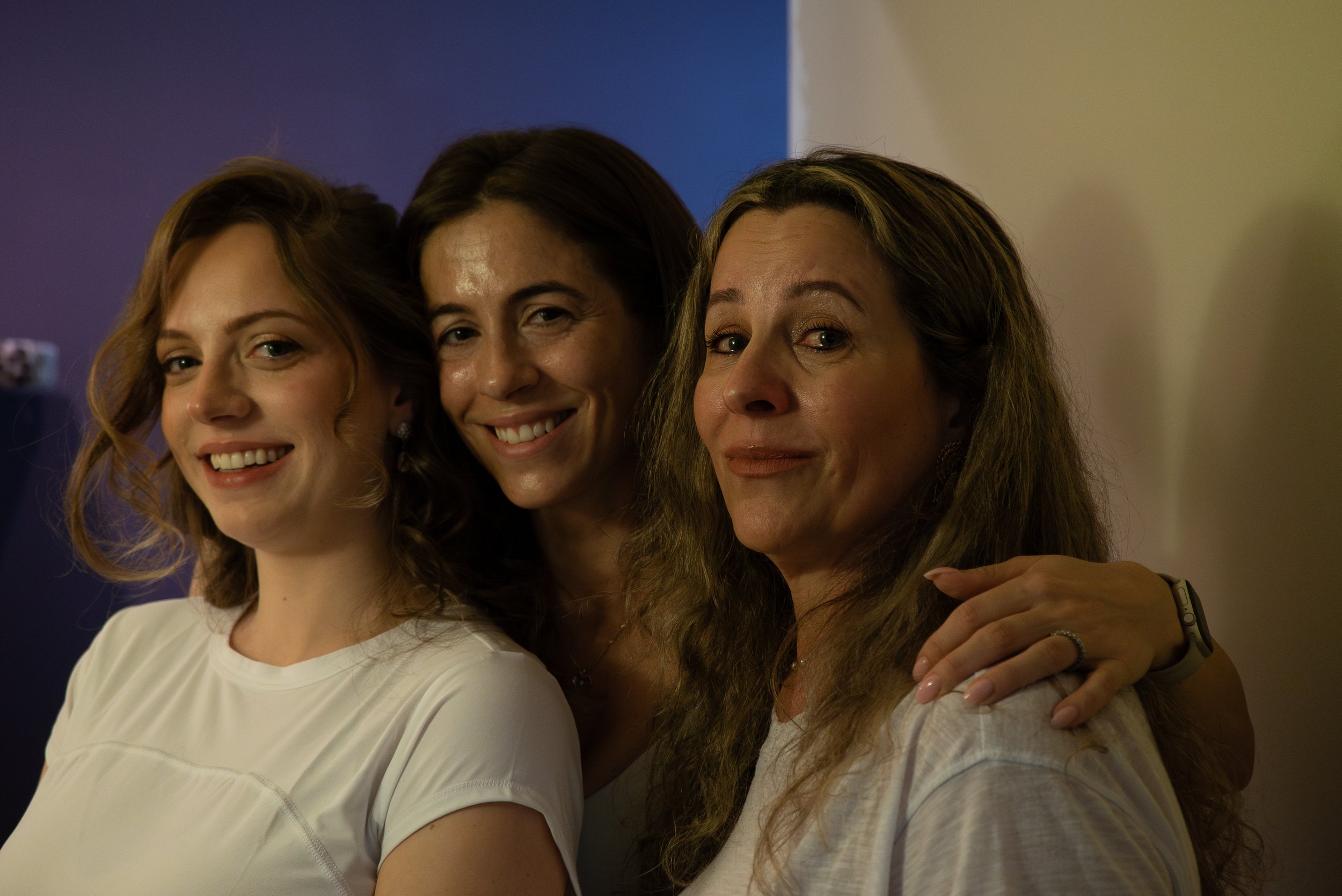 Three women smiling and hugging each other, standing close together indoors.