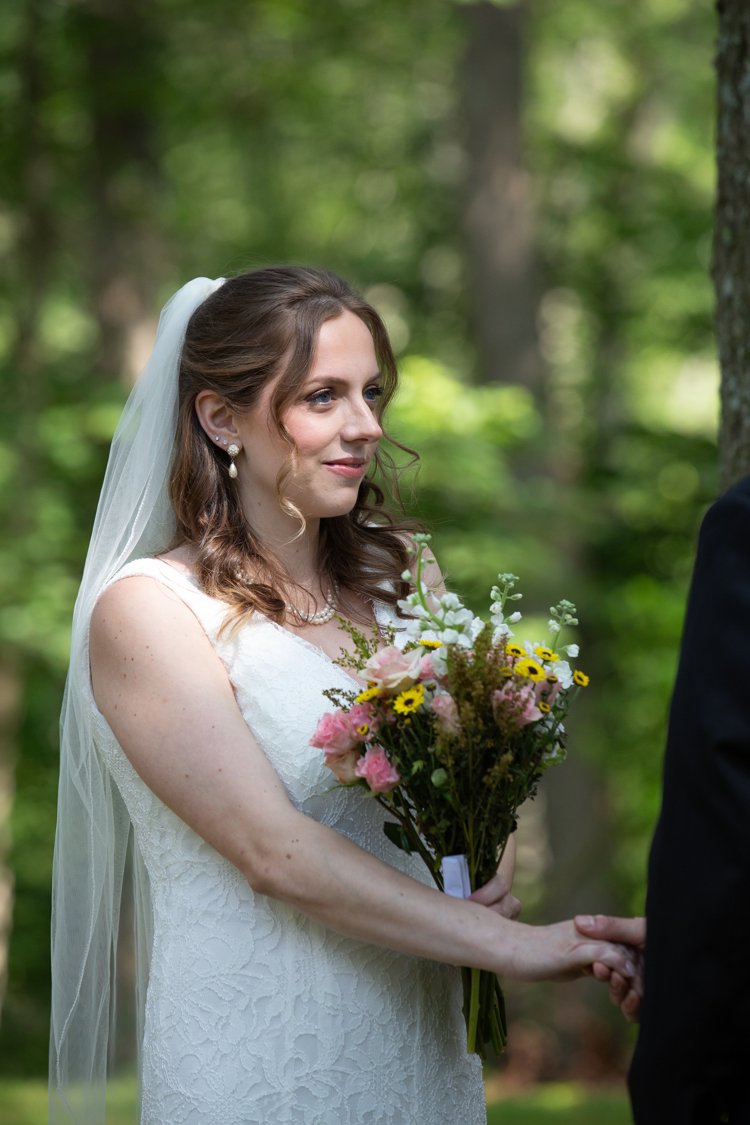 A bride with brown hair in loose curls, wearing a white lace wedding dress, veil, pearl earrings, and a pearl necklace, holding a bouquet of pink, yellow, and white flowers, shaking hands with a man in a black suit outdoors in a green forest.