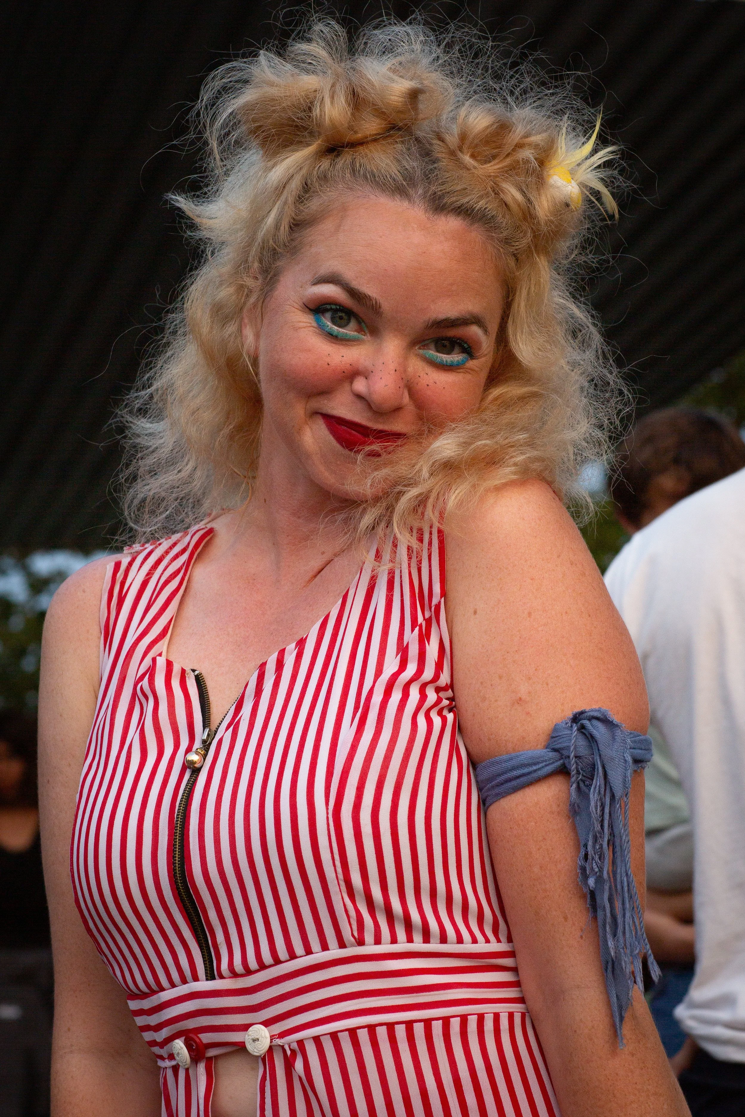 A woman with curly blonde hair styled in two buns, wearing a red and white striped sleeveless top with a zipper and buttons, along with a blue fabric armband on her arm. She has bold makeup with blue eyeshadow, freckles, and bright red lipstick, and 