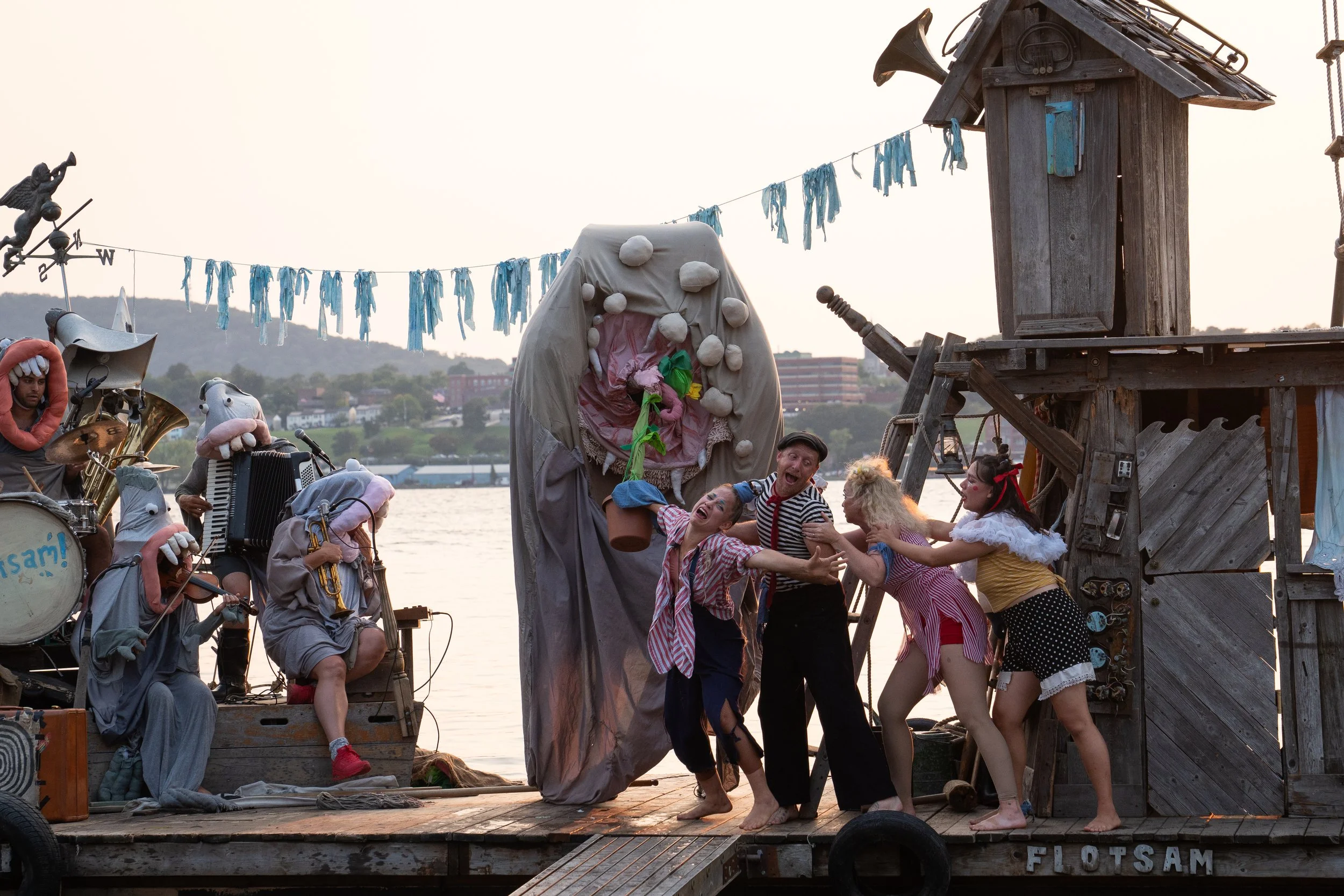 Theatrical performance on a wooden dock by a river featuring actors in colorful costumes, some wearing animal masks and playing musical instruments, with a large puppet shark centerpiece and a rustic wooden building on the right.
