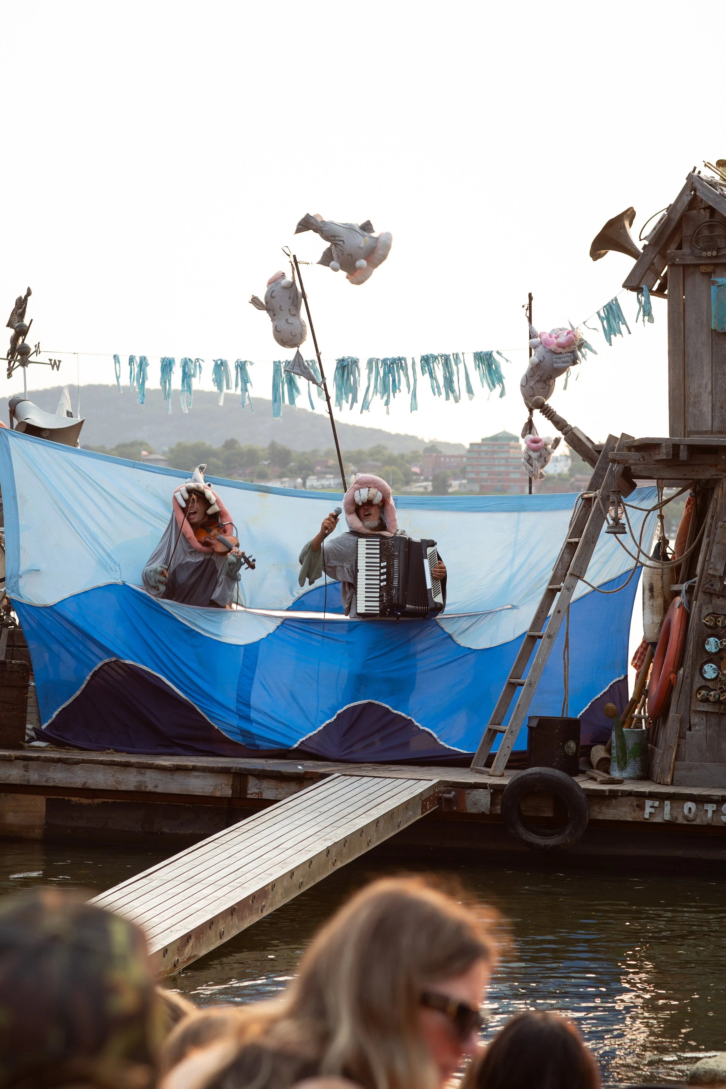 People in elephant costumes performing on a floating platform on the water, with a backdrop of hills and buildings, during sunset.