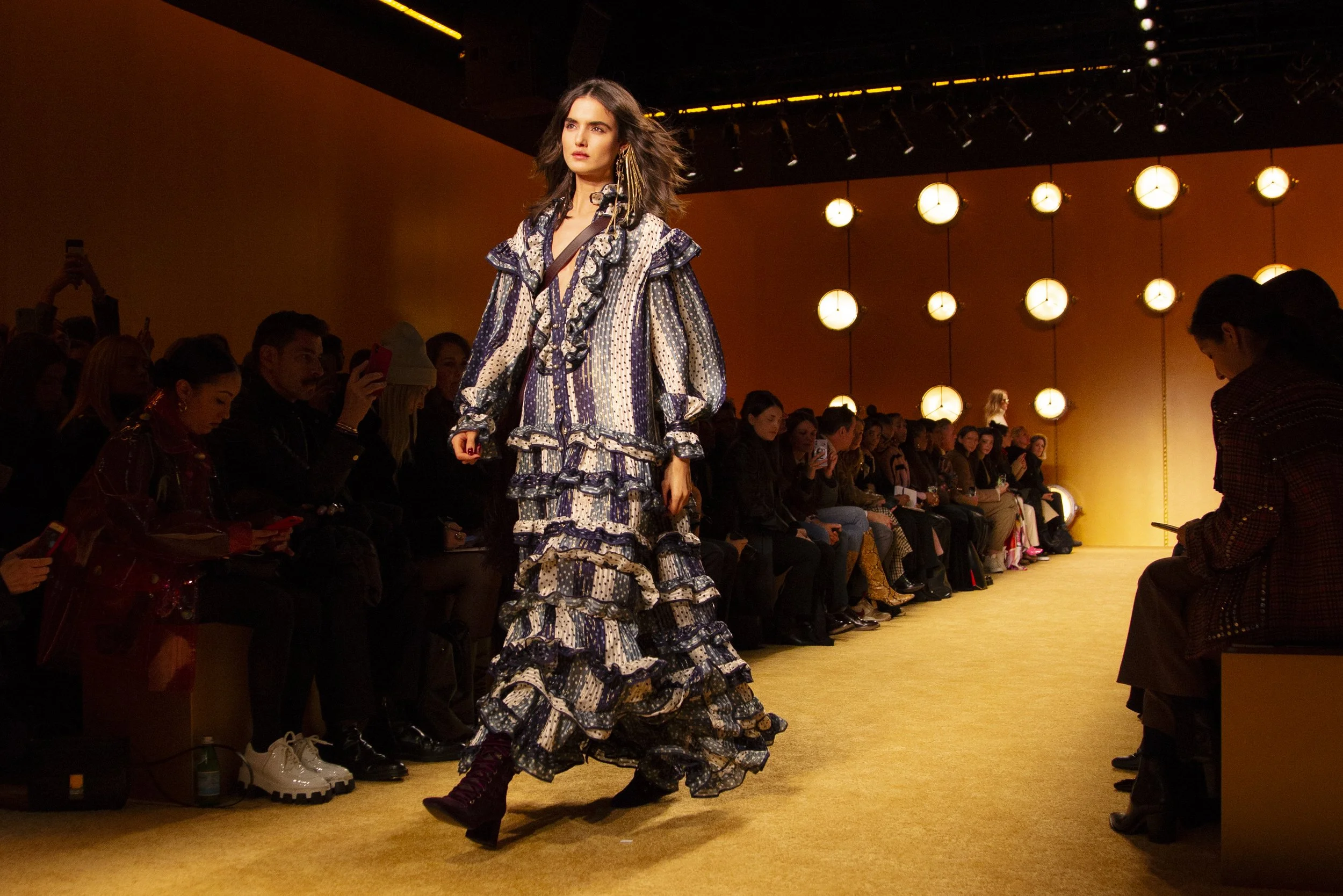 A model walks down the runway during a fashion show, wearing a long, ruffled dress with a floral pattern, while spectators sit on both sides watching and taking photos.