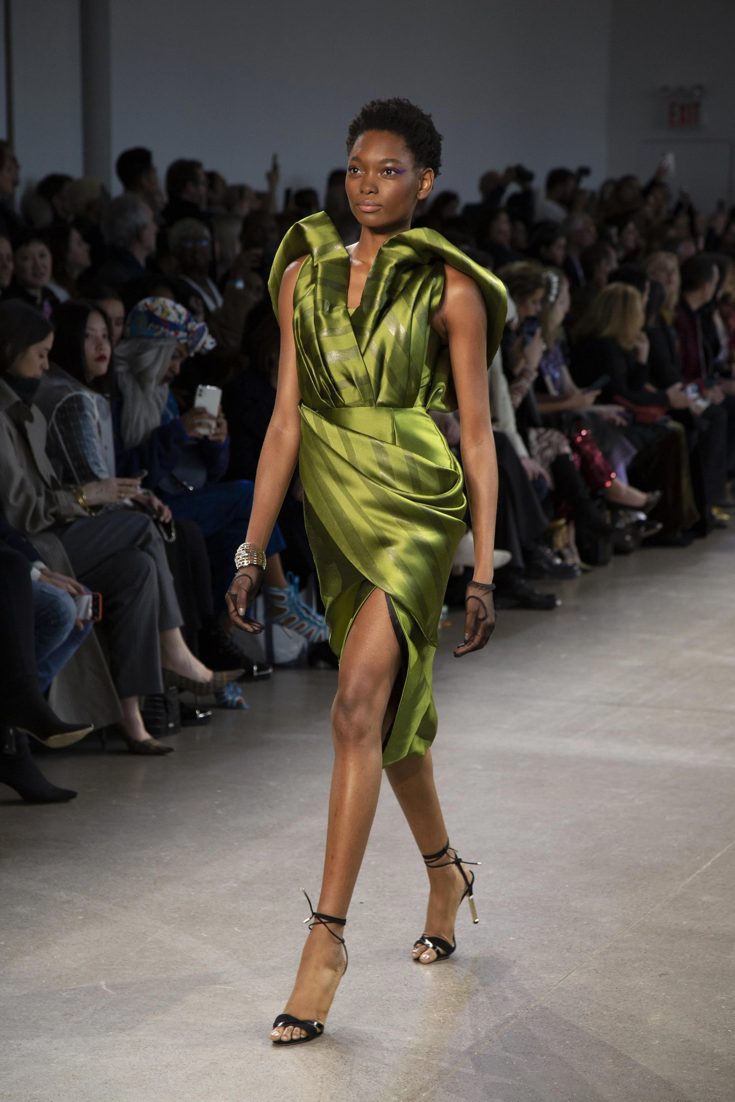 A model walks down the runway at a fashion show wearing a shiny green dress with an asymmetrical slit, paired with high-heeled sandals and gloves, while an audience watches. New York Fashion Week