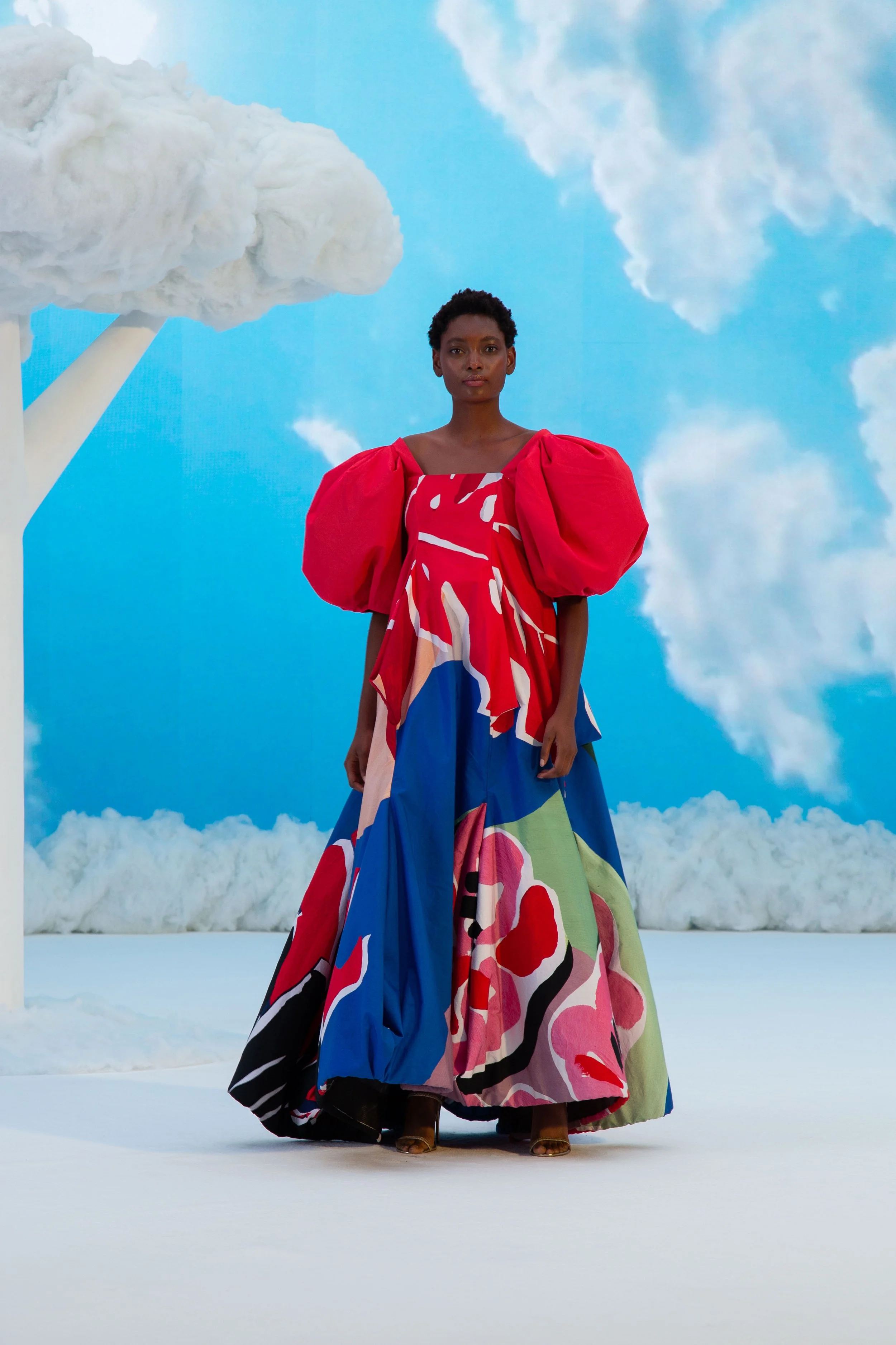 A woman wearing a colorful, abstract-designed gown with red puffy sleeves, standing on a white floor in front of a blue sky backdrop with clouds and a cotton candy-like cloud formation on the left.