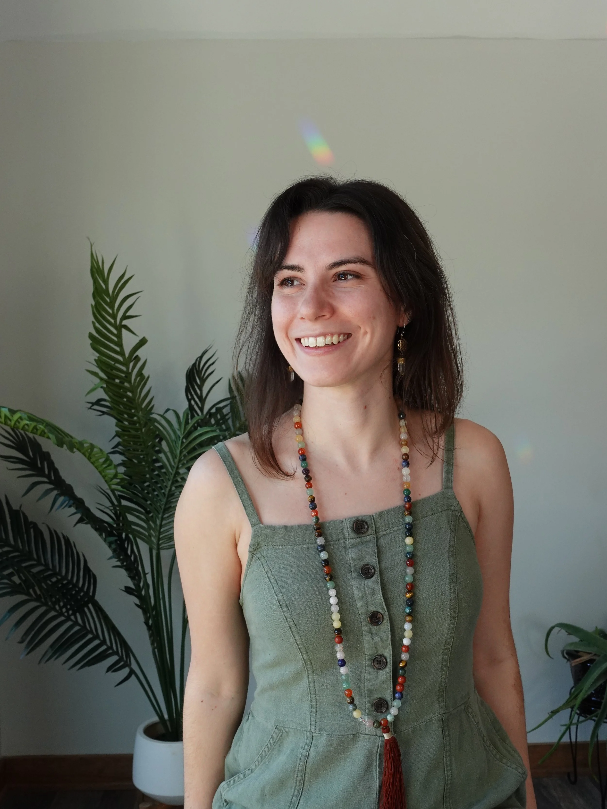 A woman with shoulder-length dark hair, smiling and looking to the side, wearing a green sleeveless button-up dress with shoulder straps, a colorful beaded necklace with a tassel, and earrings, standing near potted plants indoors.
