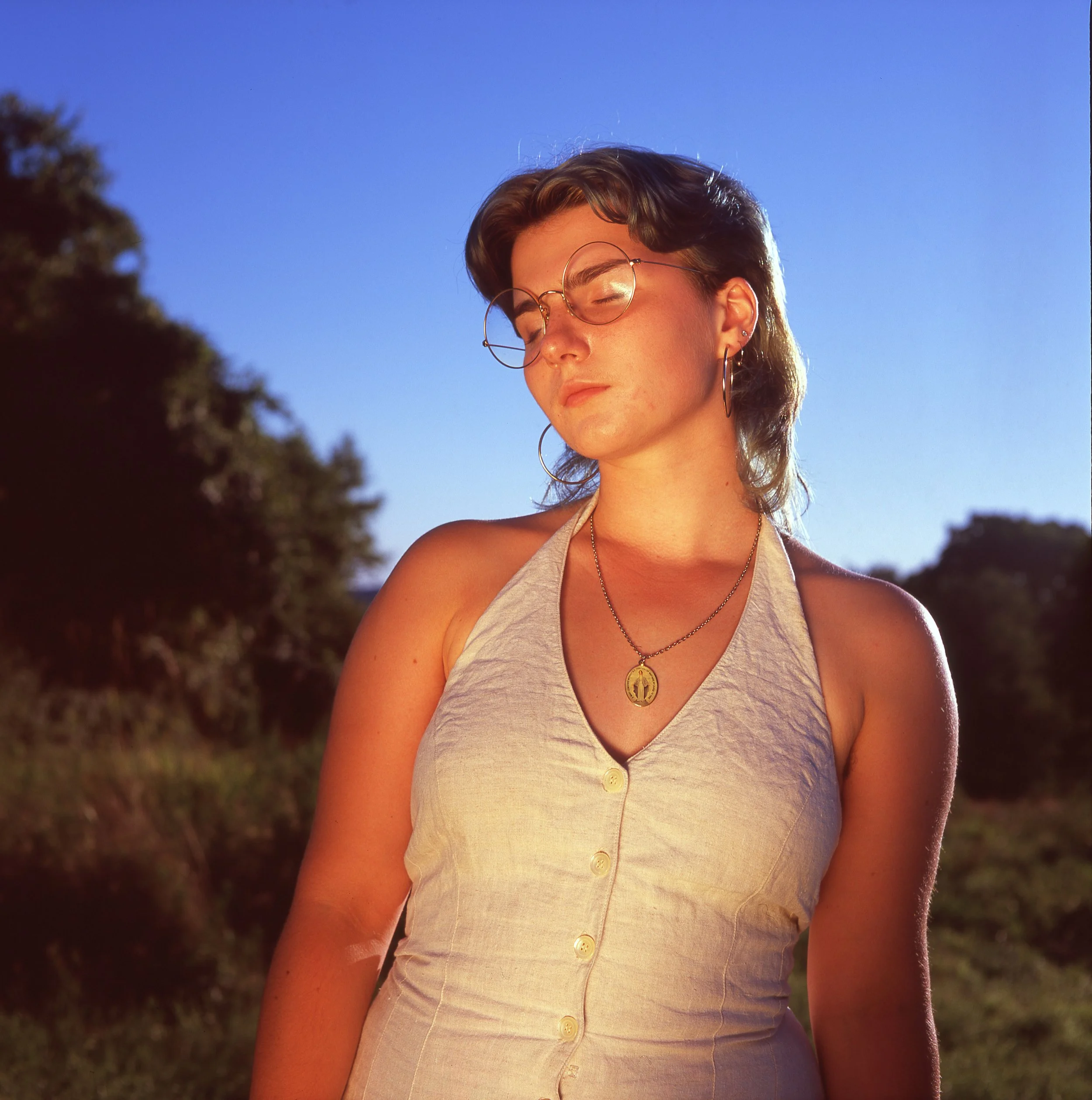 A young woman with short brown hair and glasses closes her eyes outdoors during sunset, wearing a sleeveless beige top, a gold necklace with a pendant, and hoop earrings, with trees in the background.