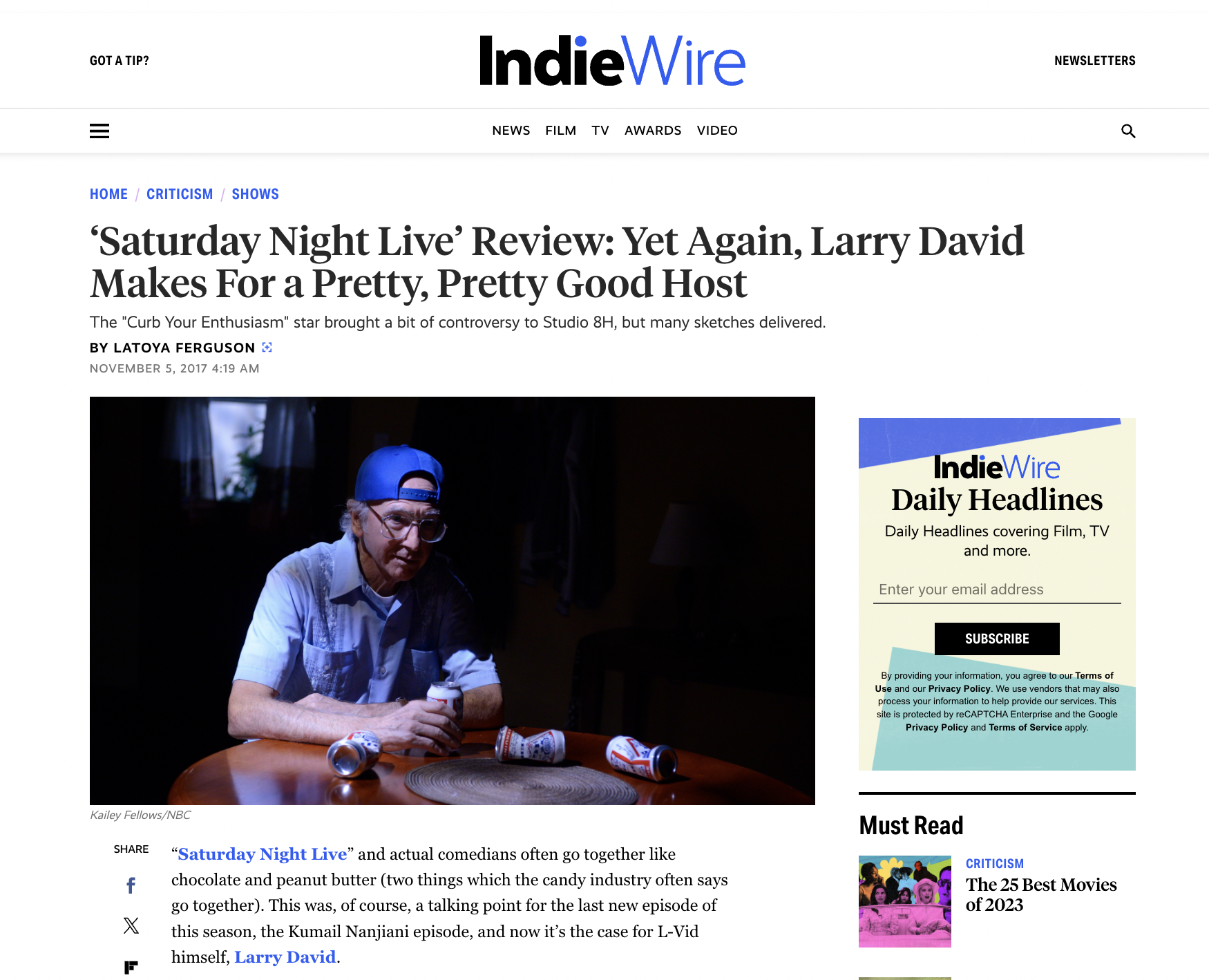 A man wearing a blue baseball cap, glasses, and a light-colored short-sleeve shirt, is sitting at a wooden table with several cans of beer, in a dimly lit room.
