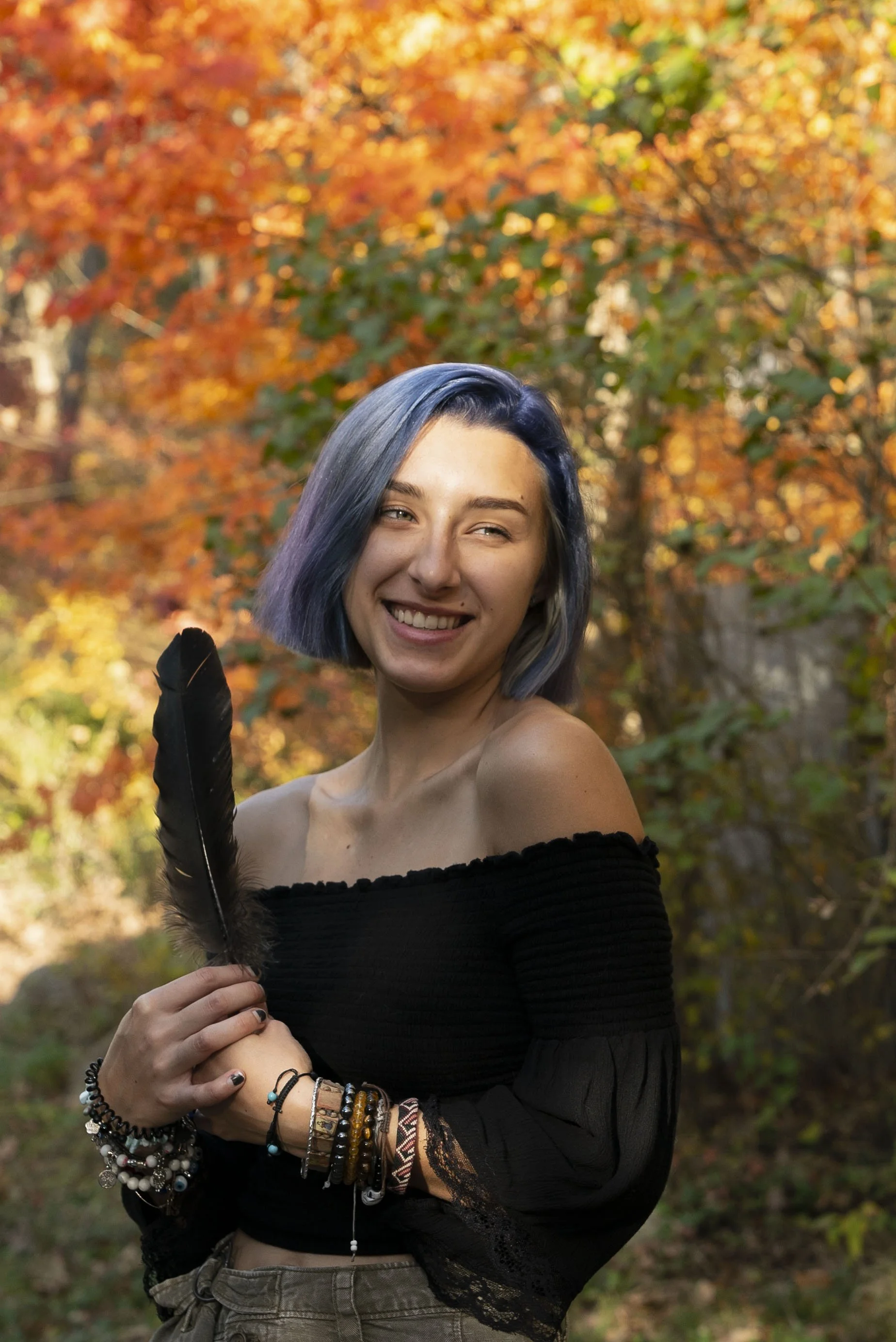 A young woman with short, blue-tinted hair smiling and holding a black feather, standing outdoors in front of fall foliage with orange and yellow leaves.