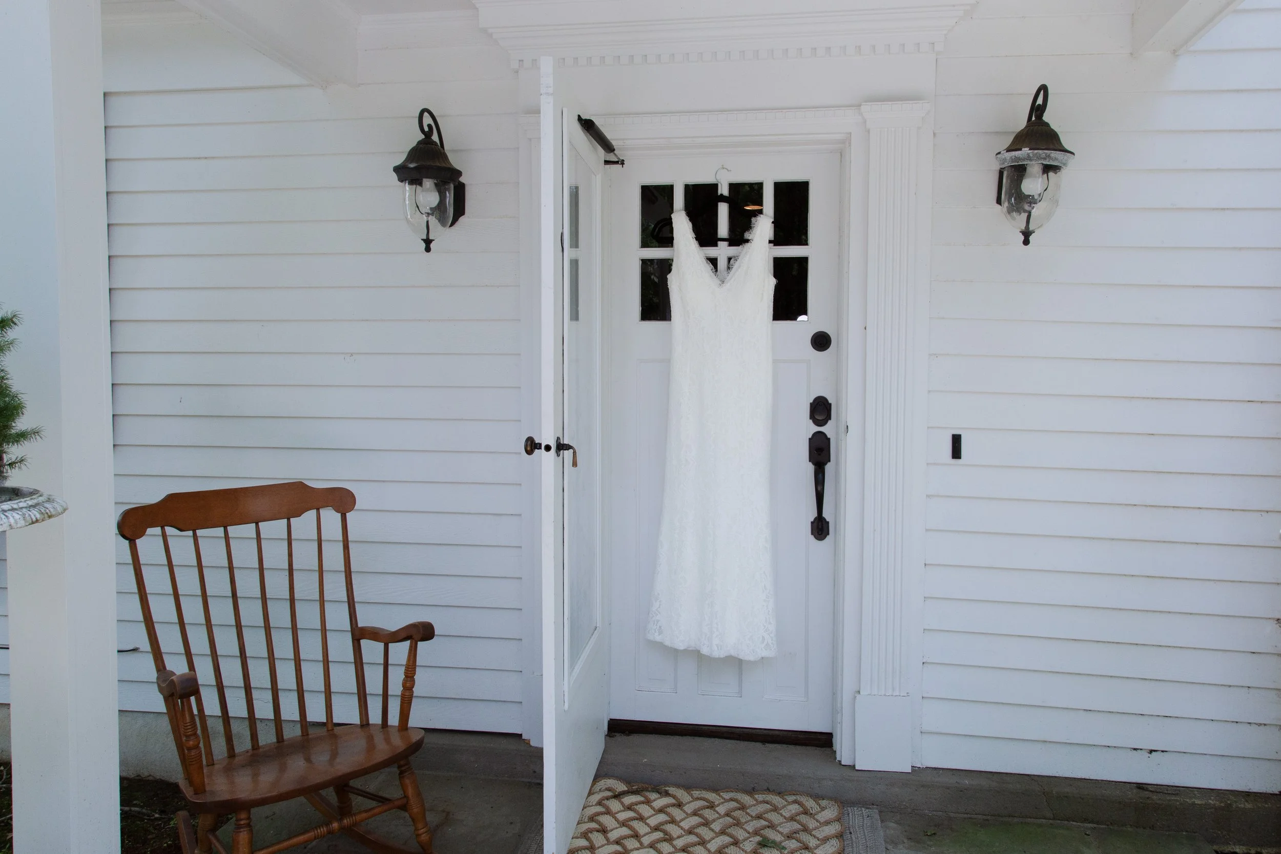 White wedding dress hanging on a door outside of a house with white siding, two black outdoor wall lanterns, a wooden chair, and a patterned doormat.