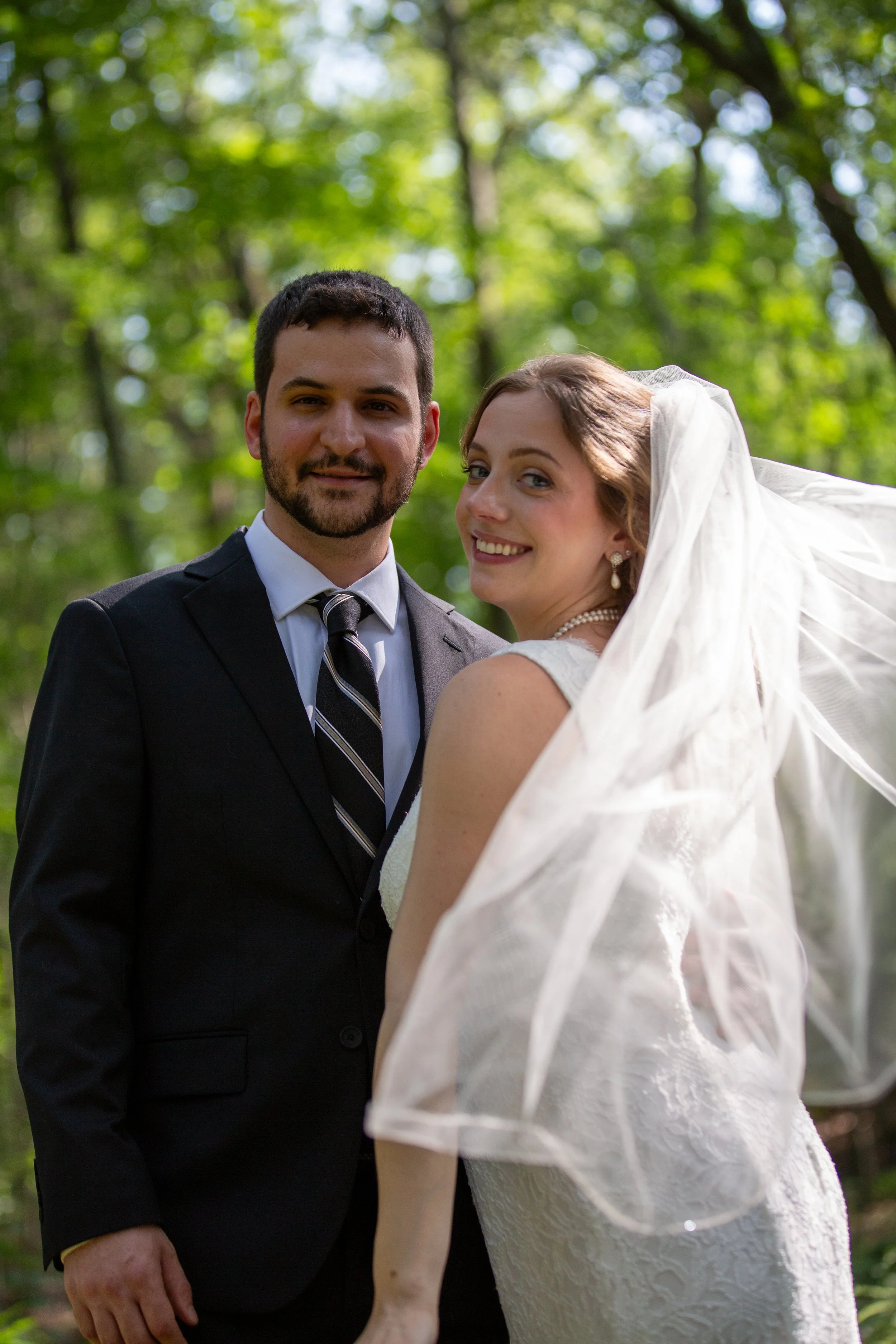 A happy bride and groom standing together outdoors in a wooded area, smiling at the camera. The bride is wearing a lace wedding dress and veil, and the groom is dressed in a black suit with a white shirt and black striped tie.