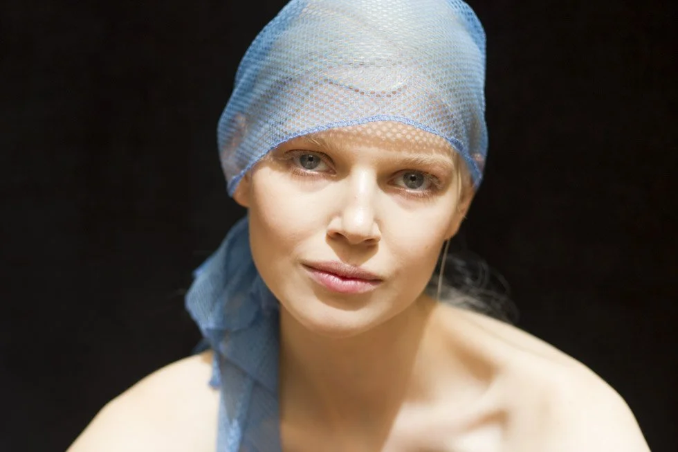 A woman with clear skin, wearing a light blue surgical cap, looking at the camera with a gentle expression, set against a black background. New York Fashion Week