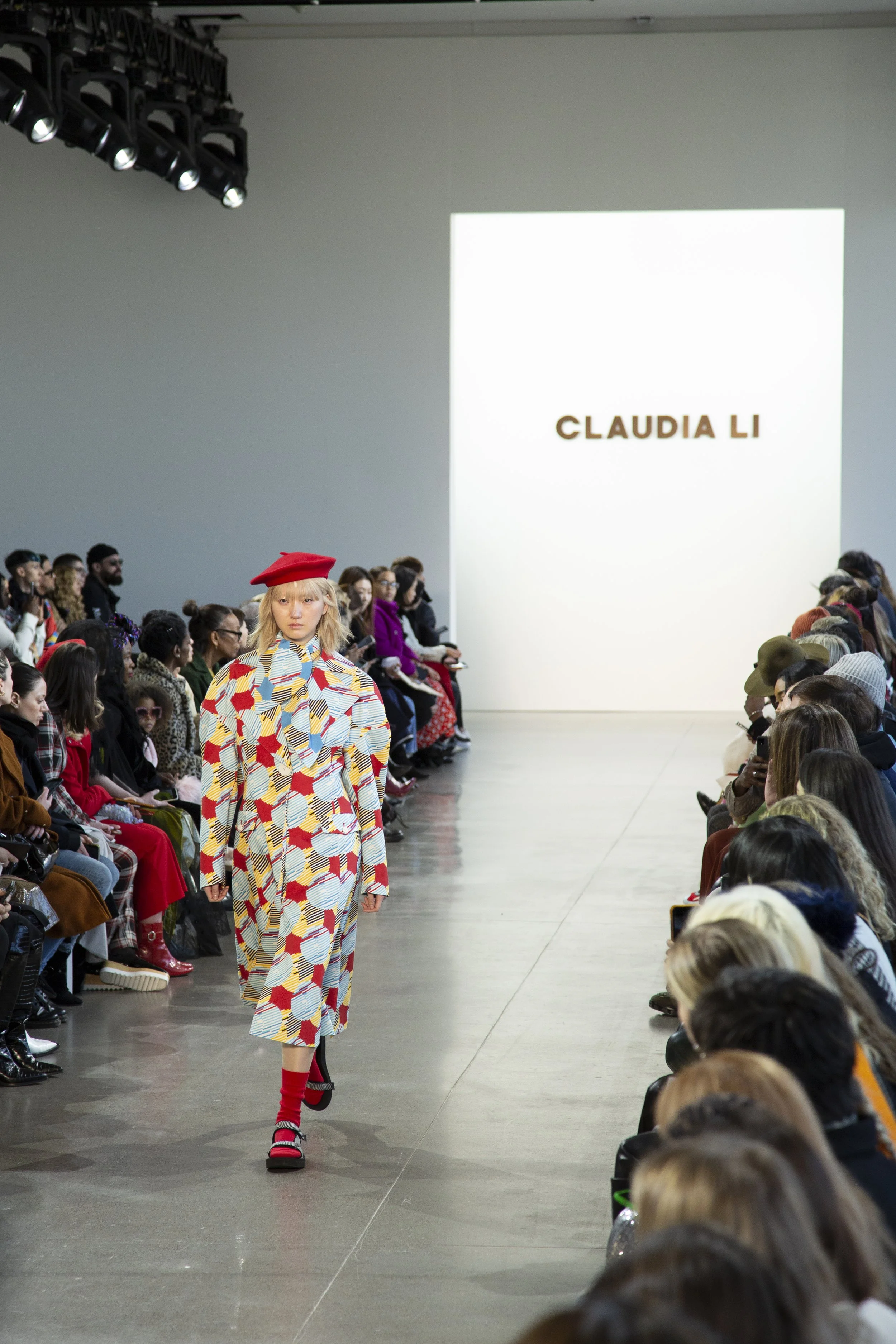 A fashion model walks down the runway wearing a colorful patterned dress with a bow at the collar, a red beret, red socks, and black shoes during a fashion show with an audience seated on both sides of the runway and a large screen displaying 'Claudi