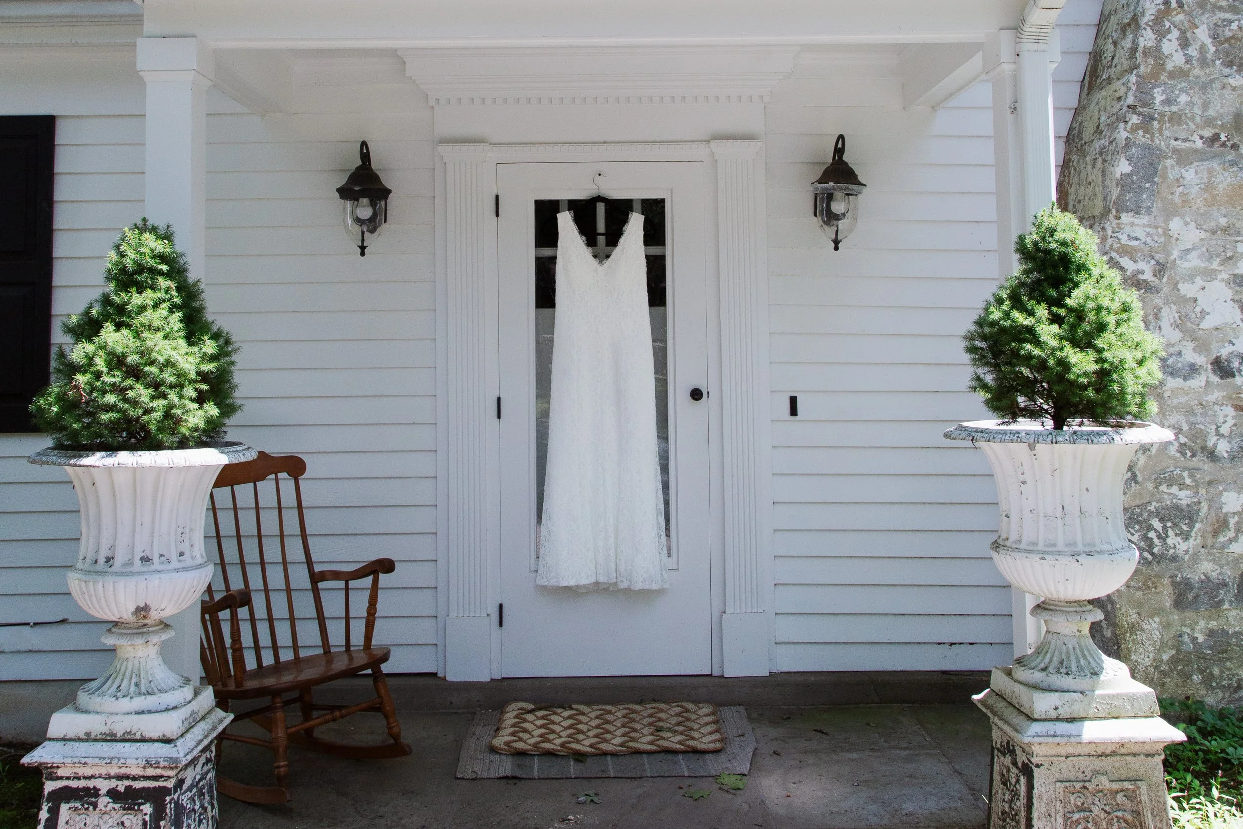 A white wedding dress hanging on a glass-paneled door on the porch of a white house, flanked by two lantern-style wall lights, with two large potted evergreen trees on either side, a wooden bench, and a small doormat on the ground.
