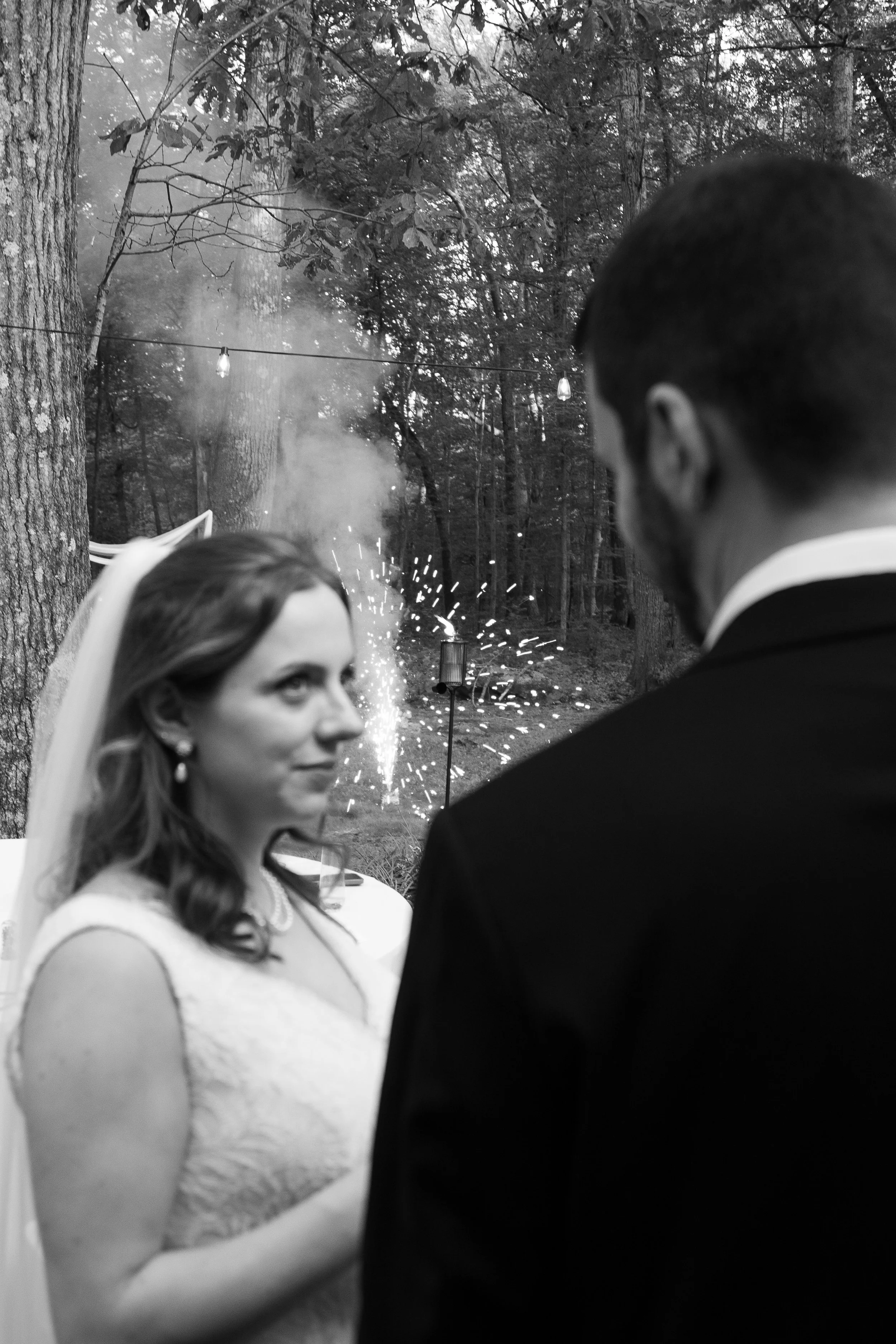 A black-and-white photo of a wedding ceremony outdoors in a wooded area with string lights hanging overhead. The bride is looking at the groom, who is facing her, with fireworks or sparklers in the background.