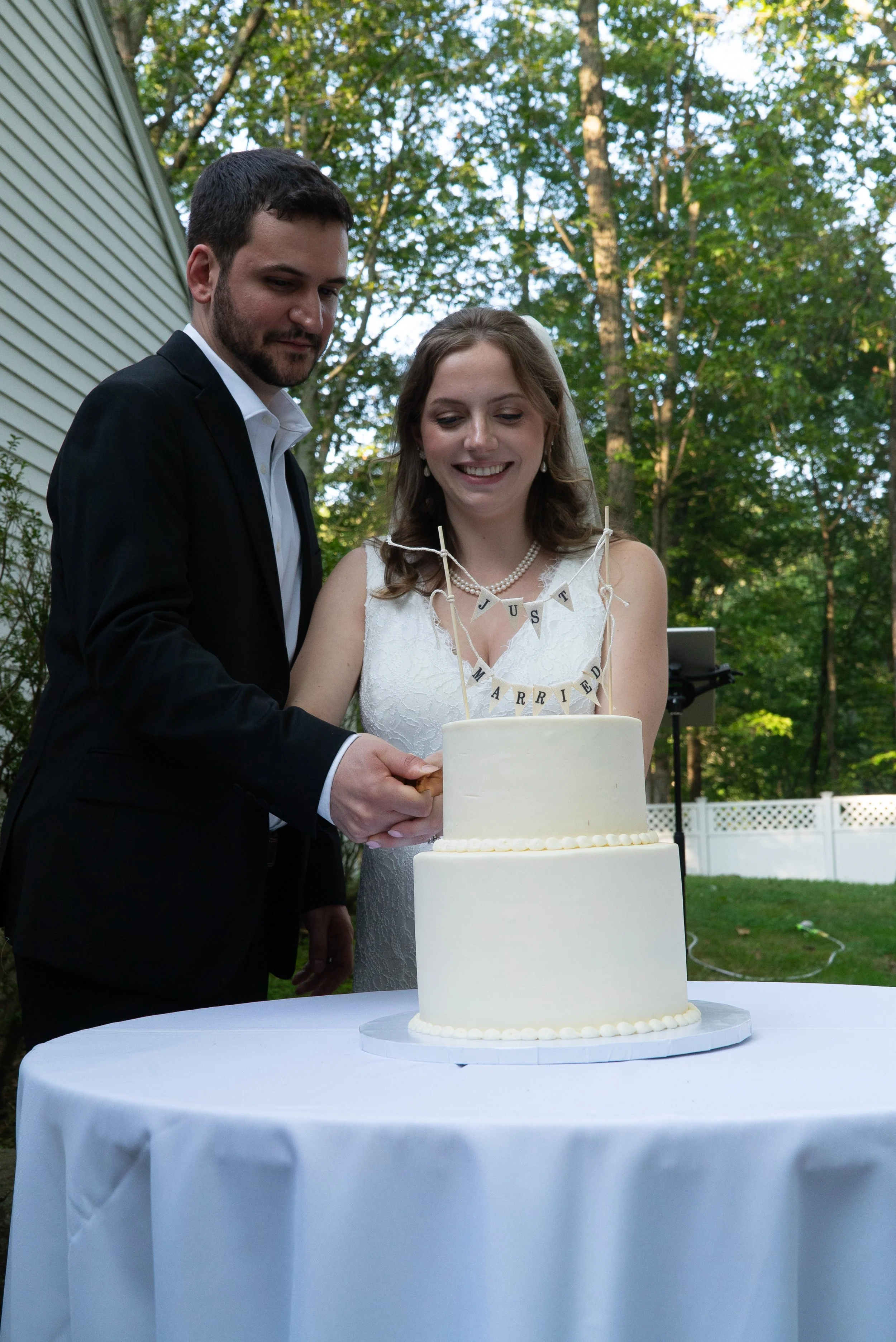 A newlywed couple cutting a wedding cake outdoors on a white table, with a 'Just Married' banner on the cake, surrounded by trees and a white fence.