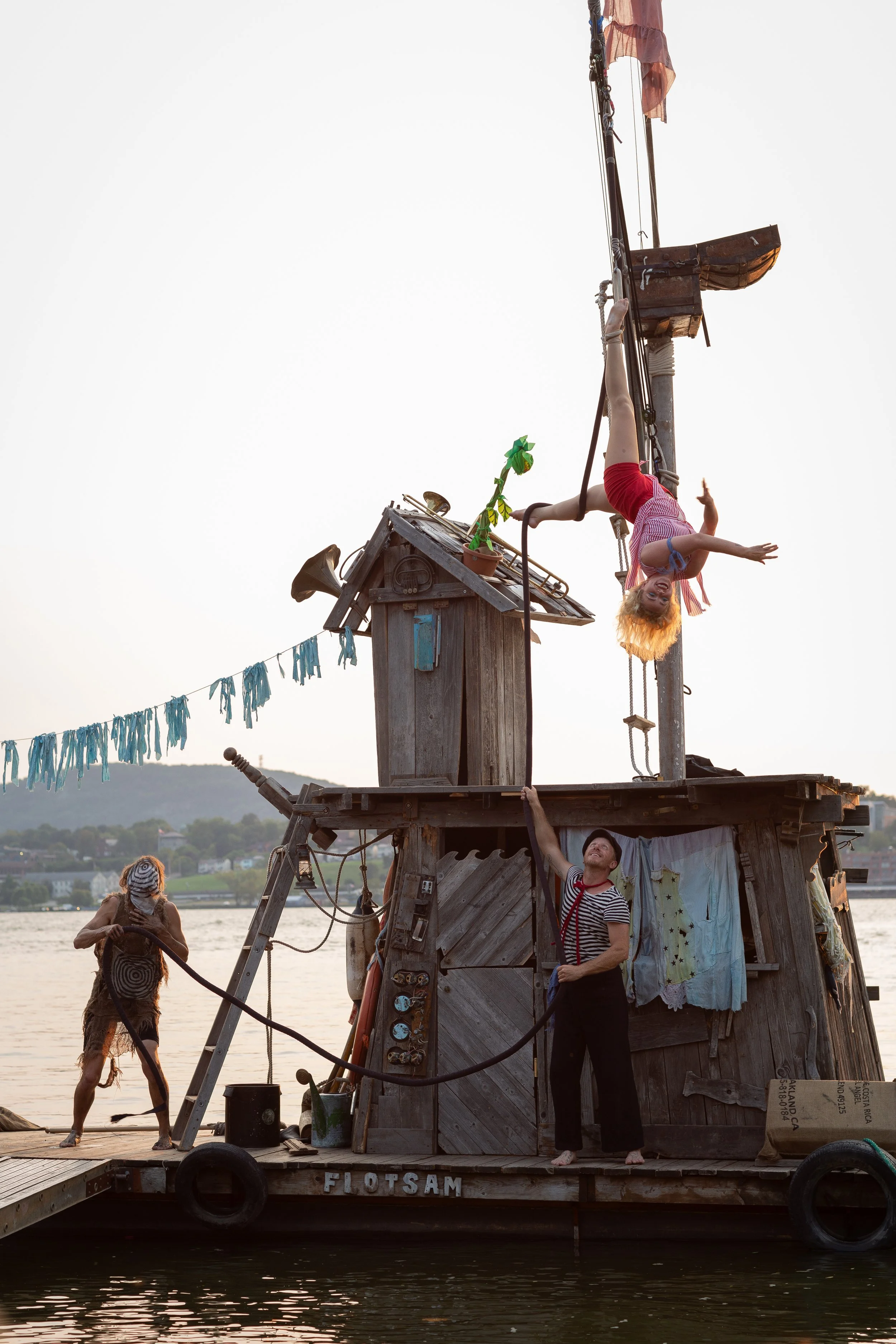 A girl is hanging upside down from a makeshift wooden house on a dock, with a man standing underneath, holding her hand. A person dressed in a Halloween costume is standing nearby, and the background features a body of water and distant hills.
