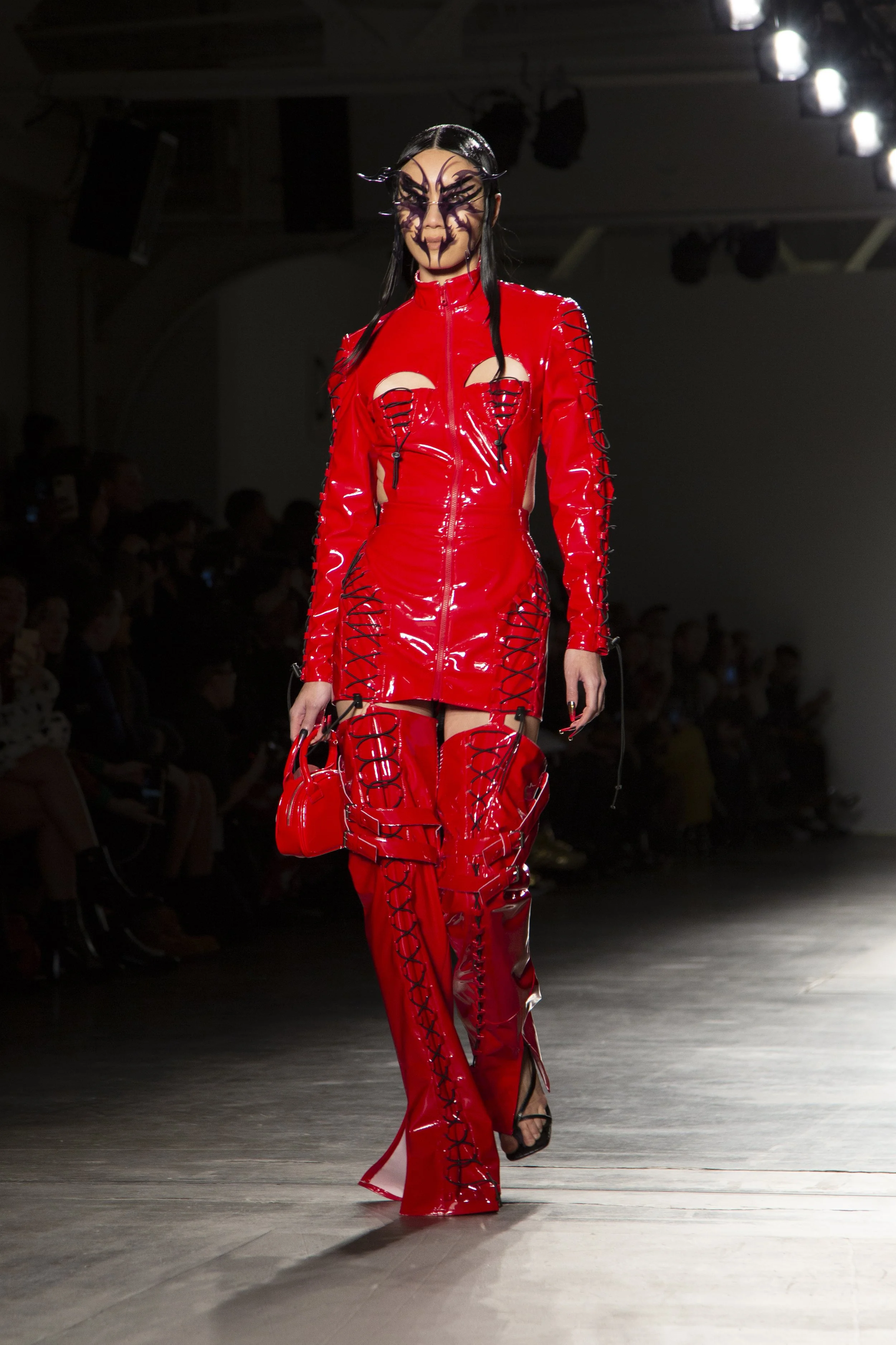 Model walking down runway in a shiny red latex outfit with black lacing details, wearing black heeled sandals, carrying a small matching red purse, and with face painted with intricate black makeup resembling a spider or insect.