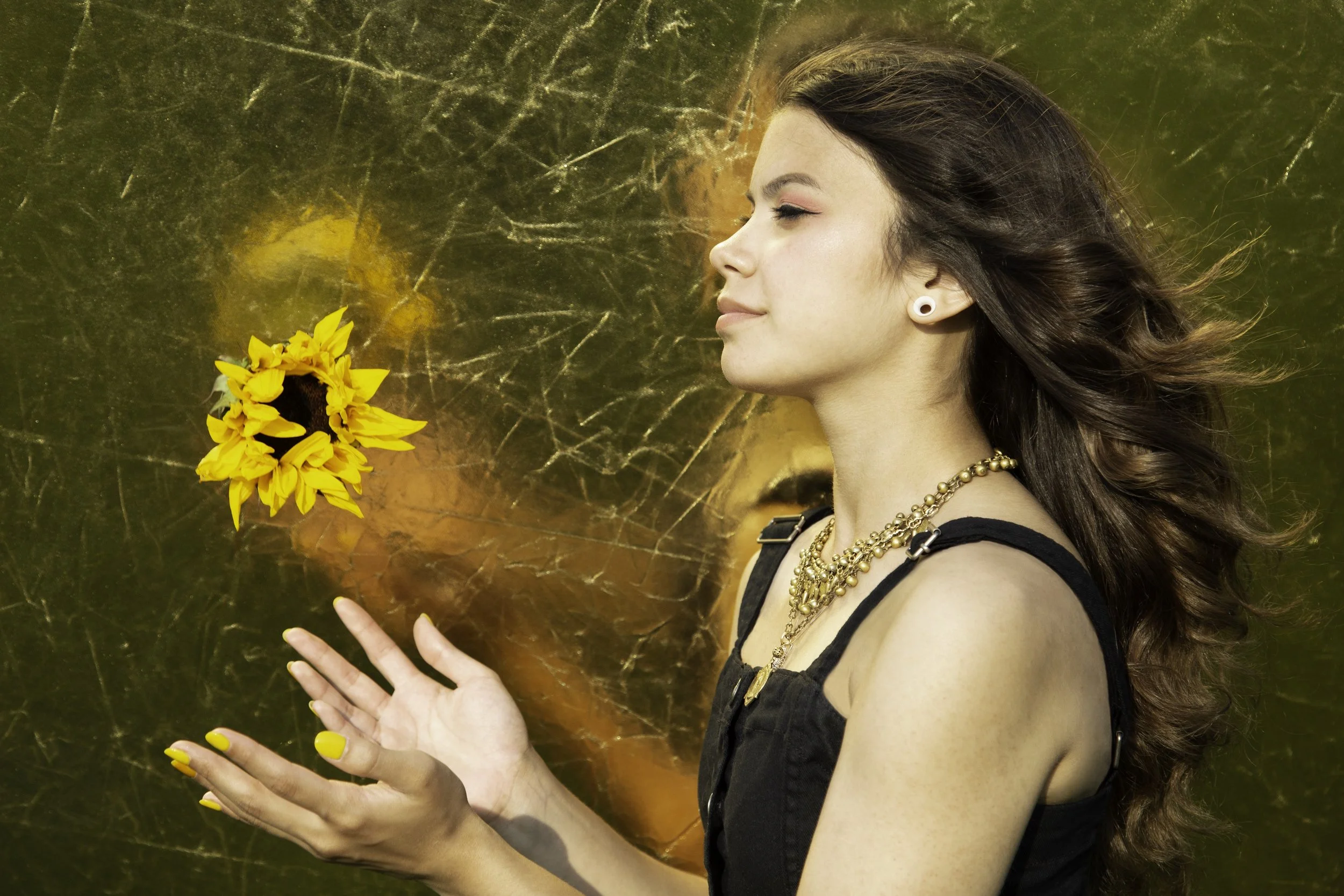 A young woman with brown, wavy hair and earrings standing against a cracked golden background, holding and looking at a sunflower.
