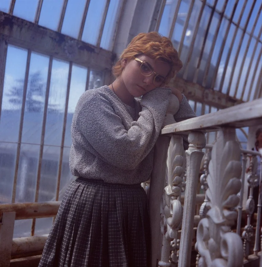 A woman with short red hair and glasses leaning on a wooden railing inside a glass greenhouse, wearing a gray sweater and a plaid skirt.