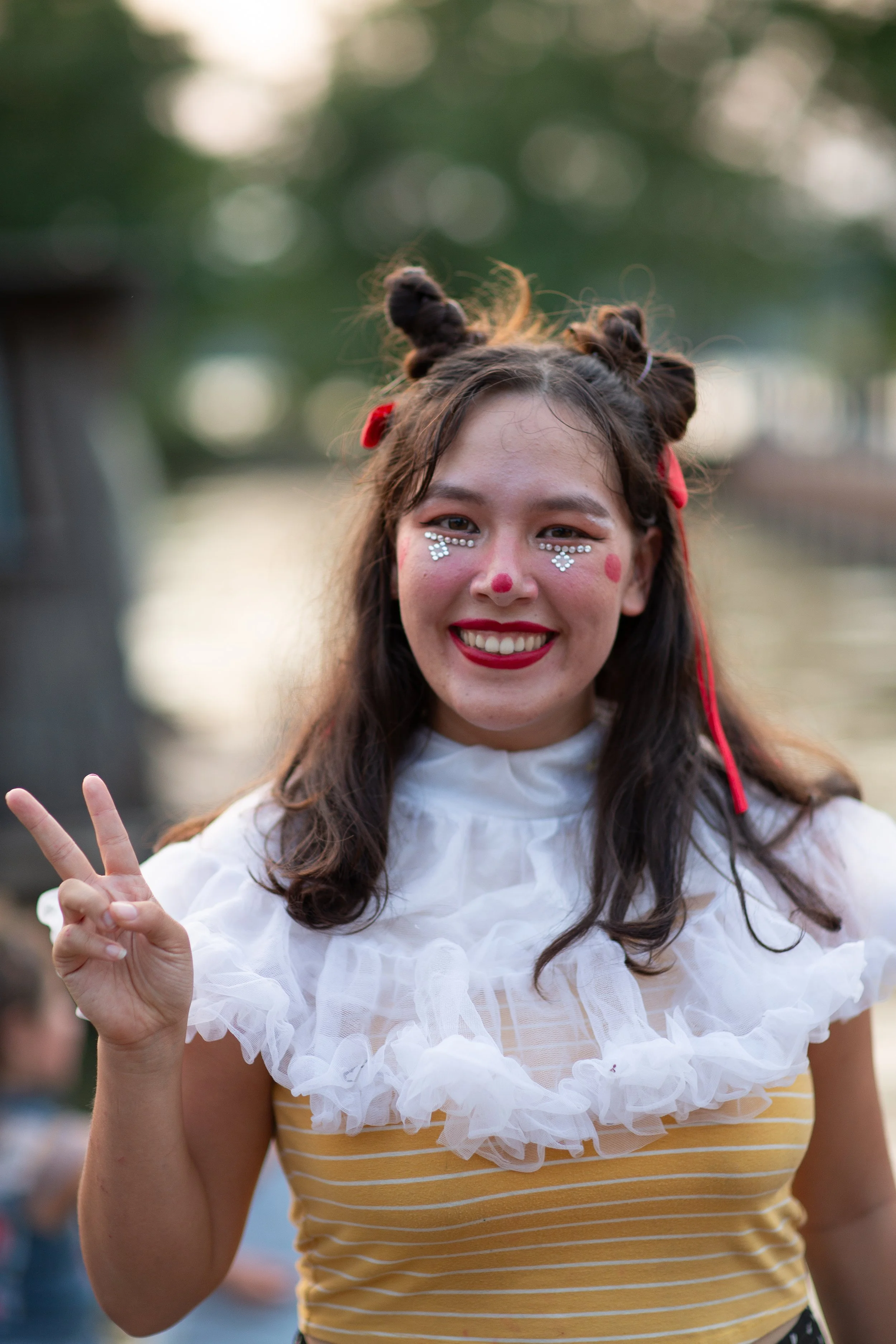 Smiling woman dressed as a clown with face paint, making a peace sign outdoors during daytime.