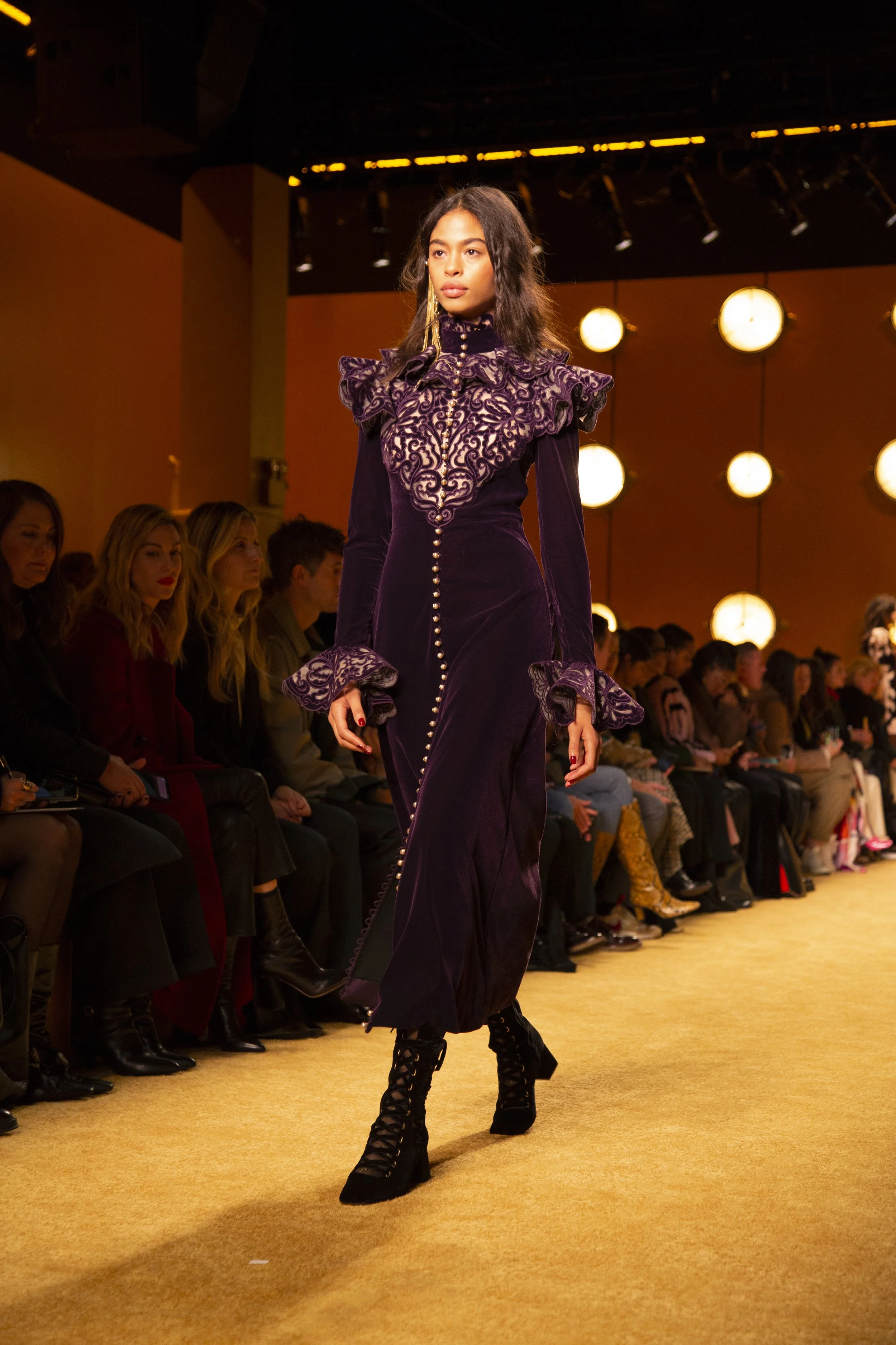 Model walking down runway in purple dress with intricate lace details and high-heeled lace-up boots, audience seated on either side, warm lighting overhead.