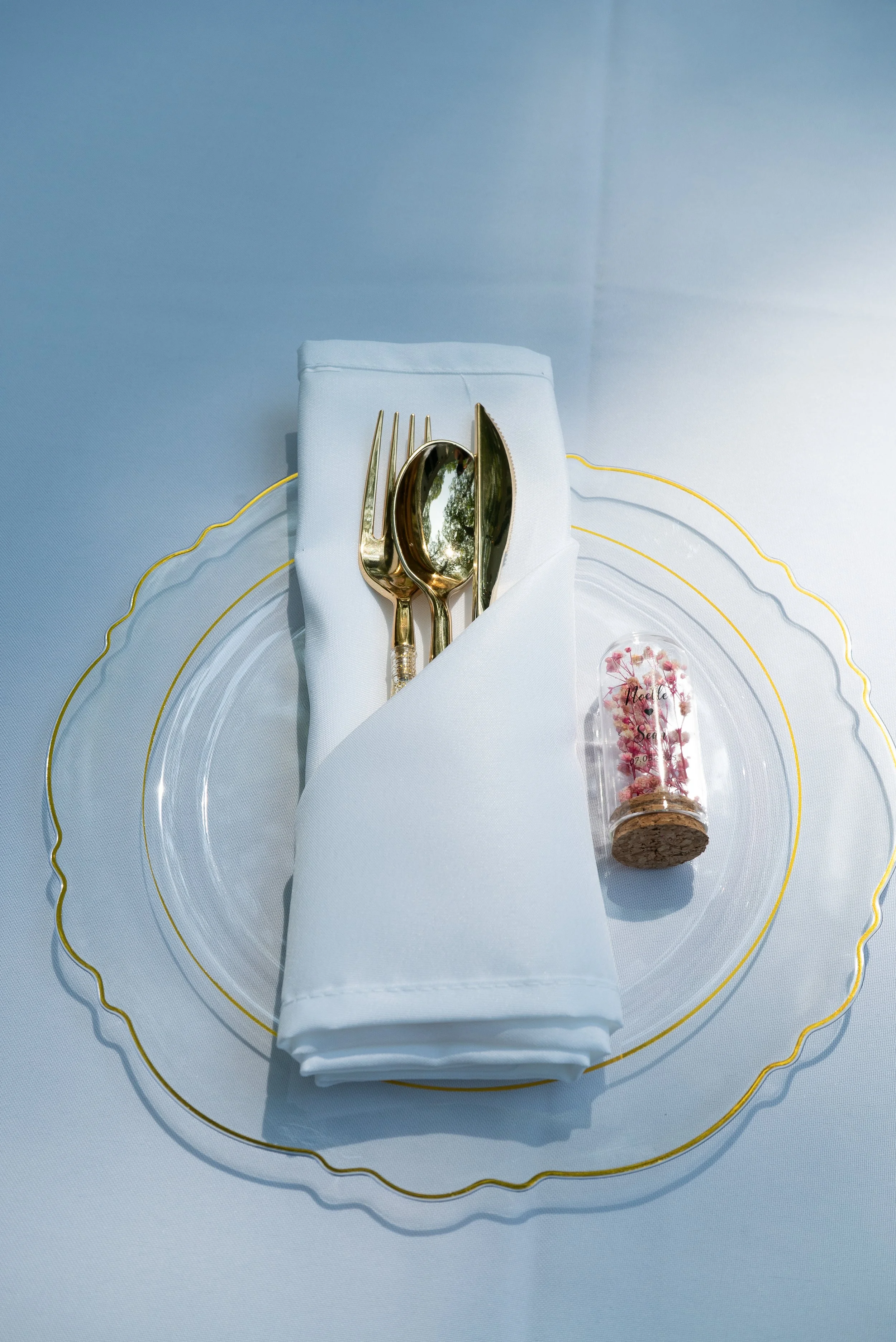A table setting with gold cutlery on a white napkin, a clear glass plate with a gold-rimmed scalloped edge, and a small glass container with pink dried flowers inside, placed on a light blue tablecloth.
