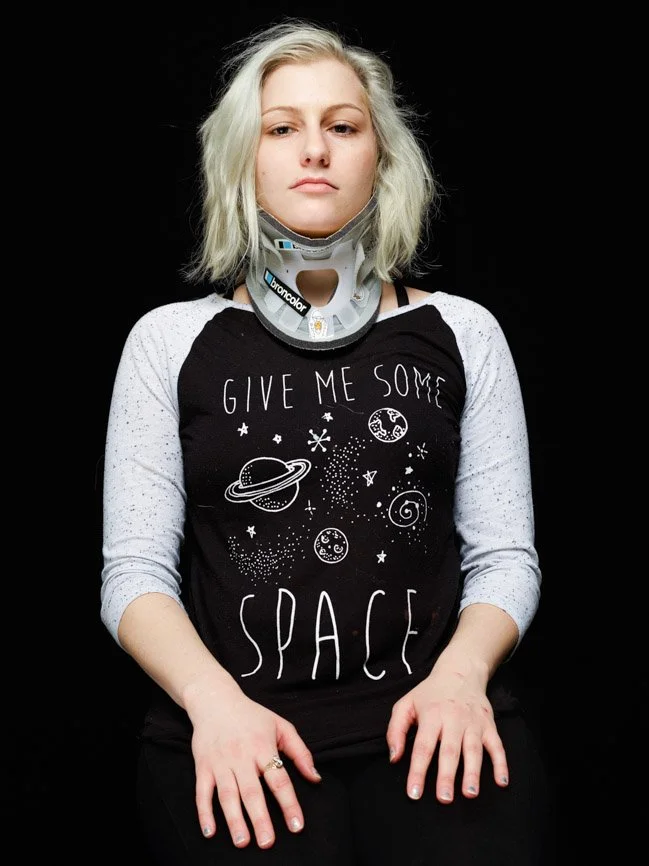 Young woman with a medical neck brace and a space-themed T-shirt against a black background.