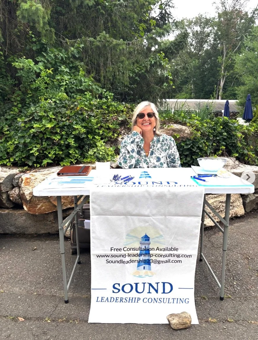 A woman sitting at a table outdoors with a banner that reads "Sound Leadership Consulting". The woman is wearing sunglasses and smiling, surrounded by green trees and bushes.