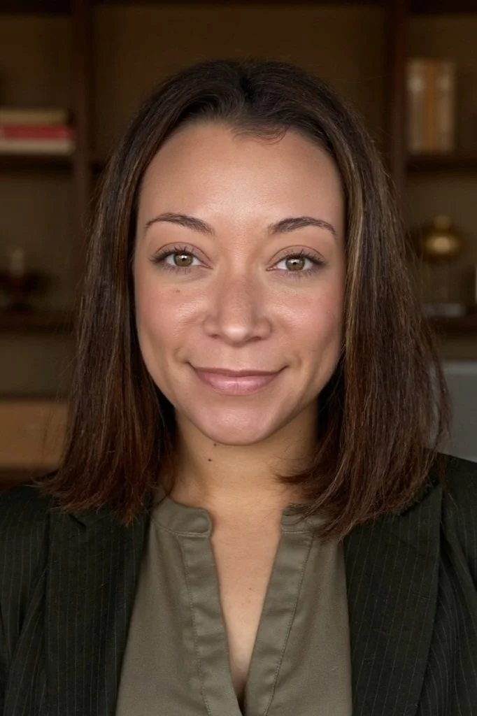 A woman with shoulder-length brown hair, light skin, and green eyes, smiling in an indoor setting with shelves and books in the background.