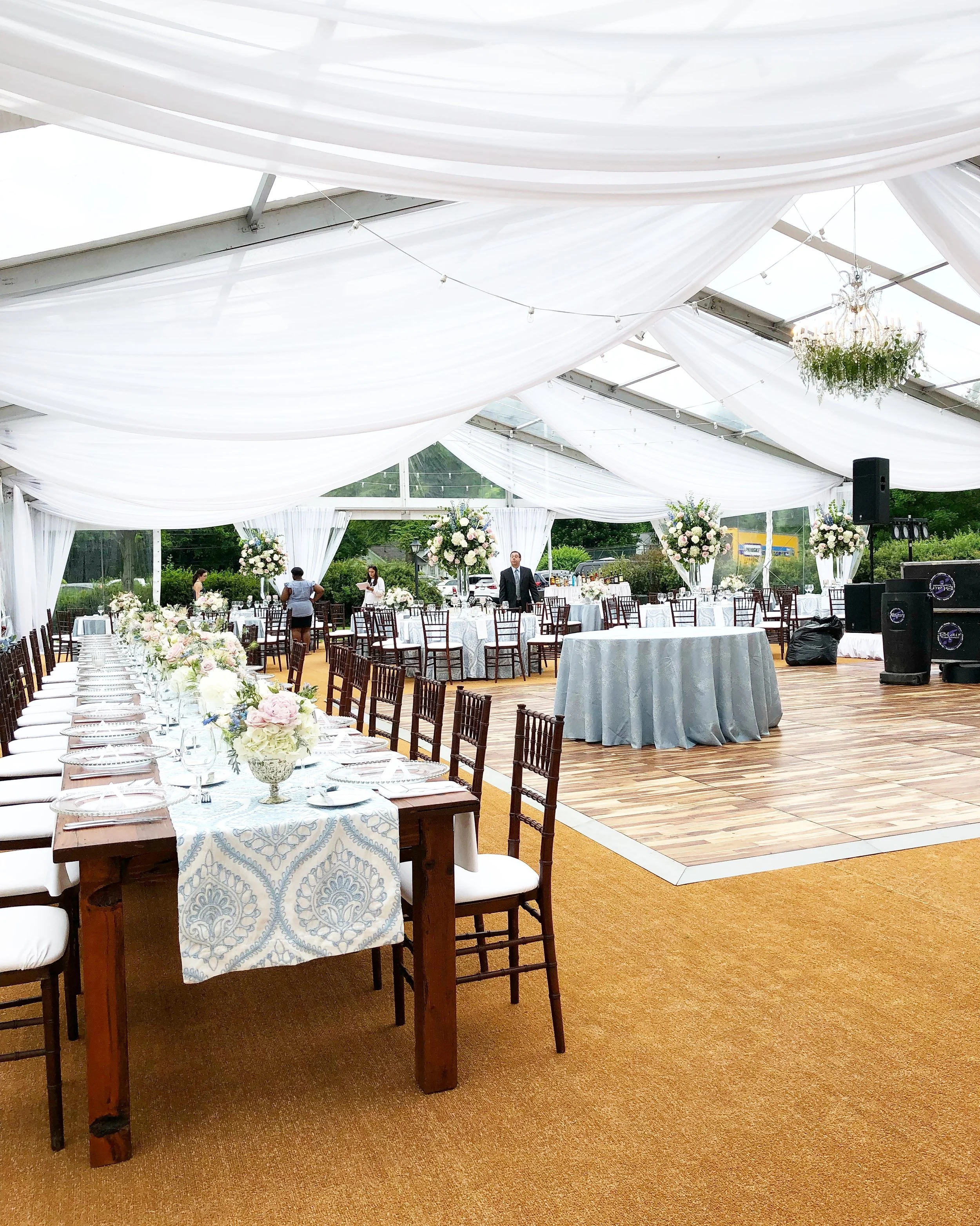 Elegant event setup inside a white tent with draped ceiling, long dining tables with floral centerpieces, and a dance floor with a gray tablecloth in the middle.
