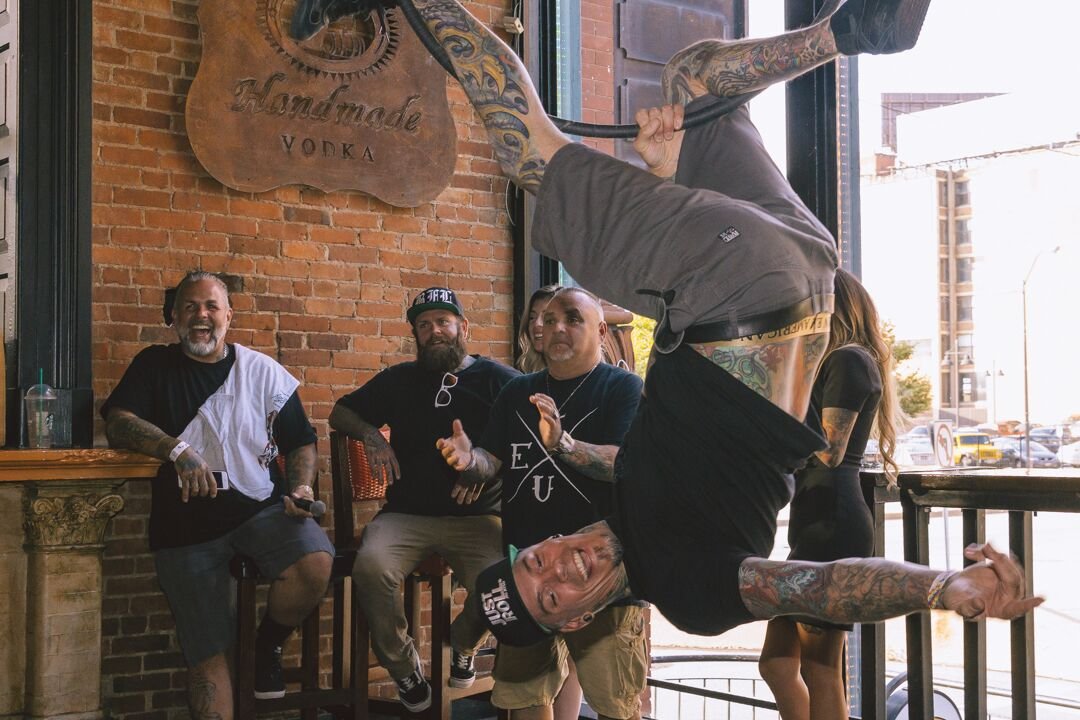 Man with tattoos hanging upside down and performing a handstand indoors, with three men watching and smiling, in a brick-walled venue with large windows.
