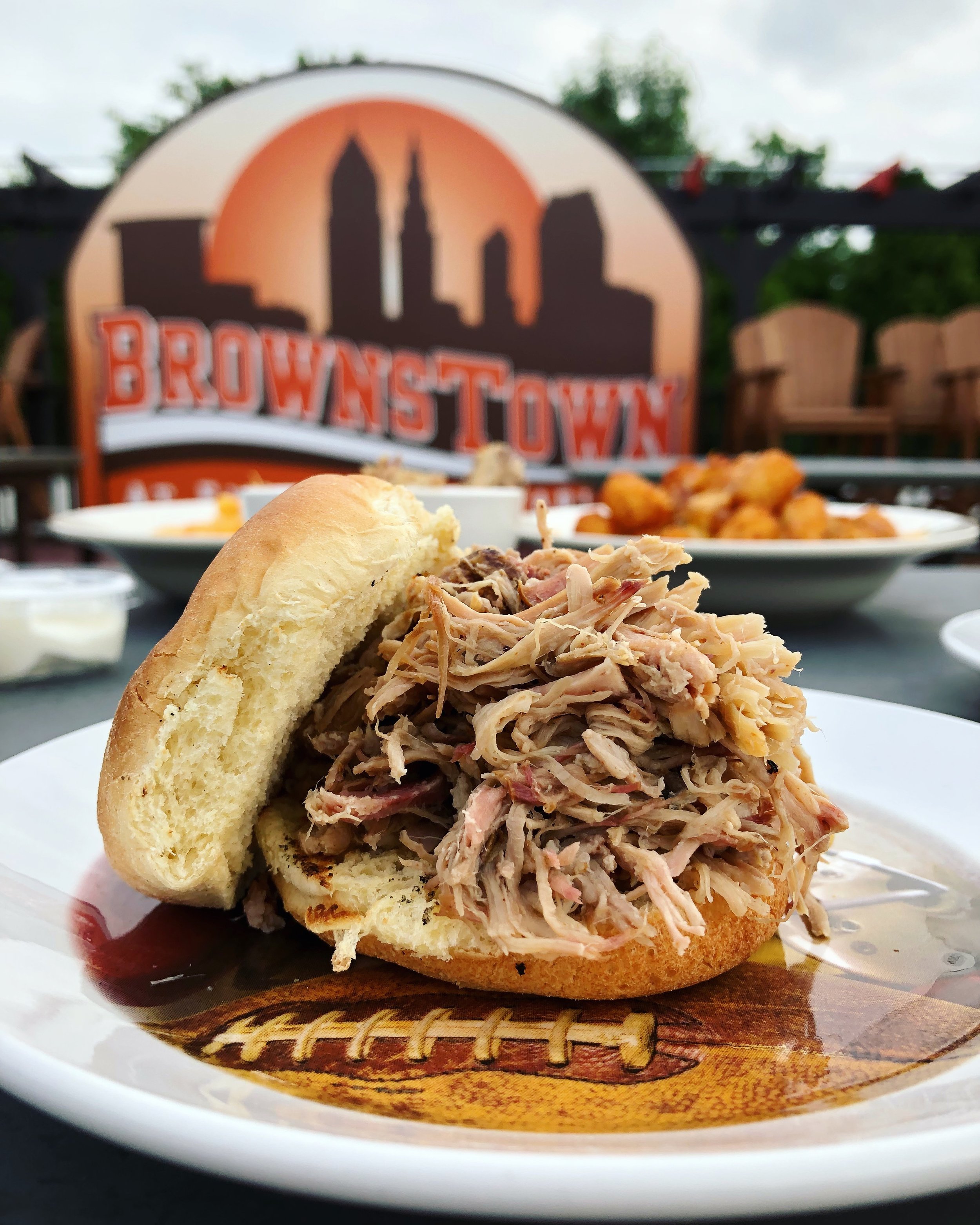 Pulled pork sandwich on a bun with barbecue sauce, set on a sports-themed plate with a brownstown sign in the background.
